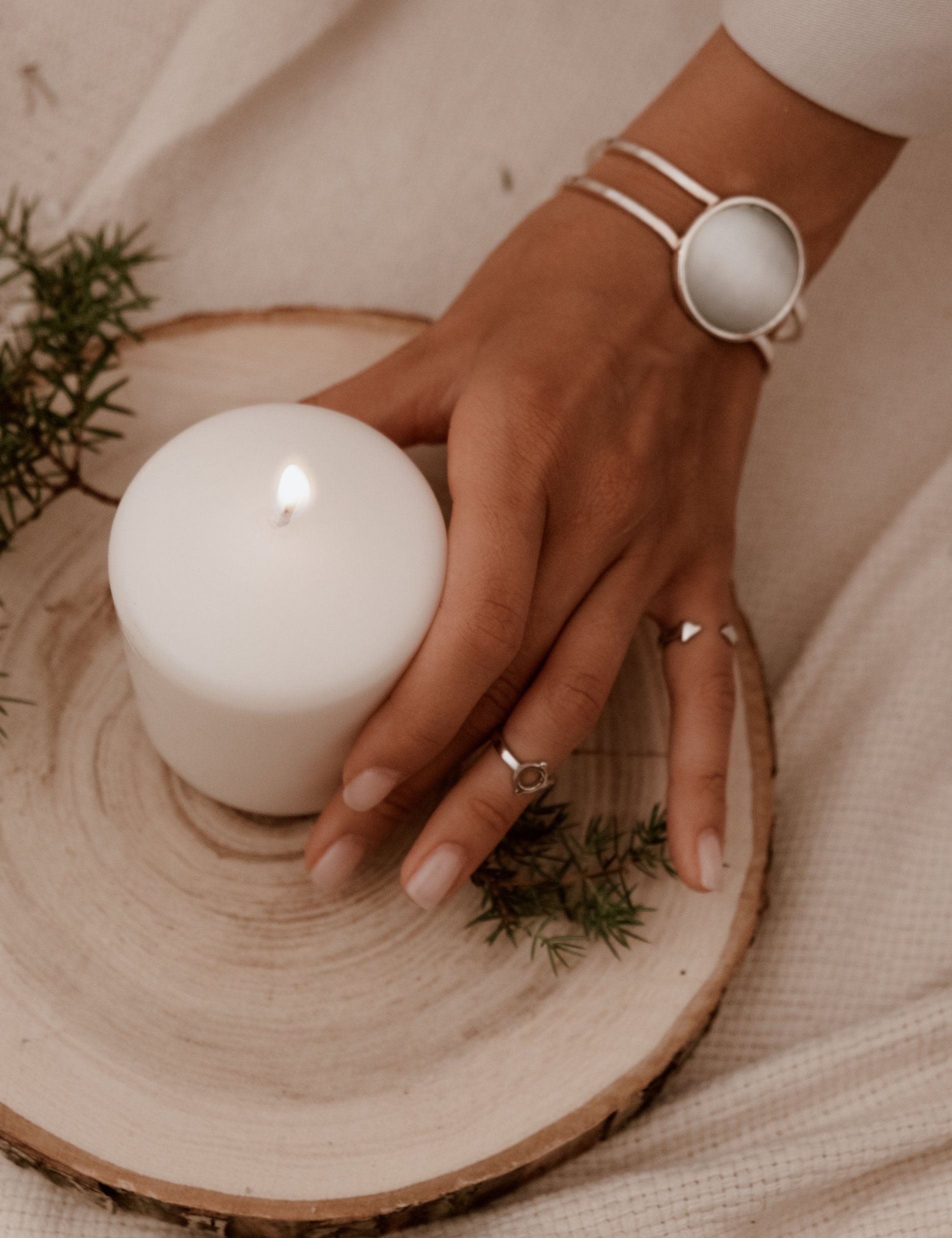 Hand with jewelry rests on a wood slice next to a lit white candle.