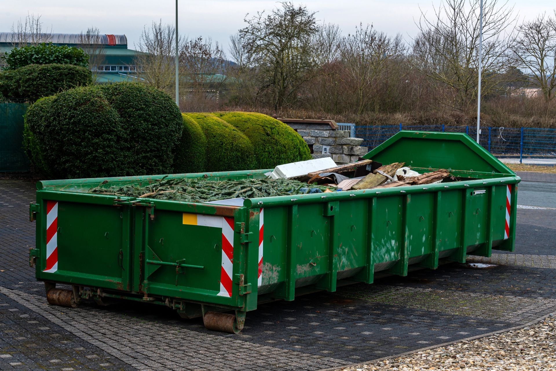 Green dumpster filled with debris, parked outside near bushes and a building.