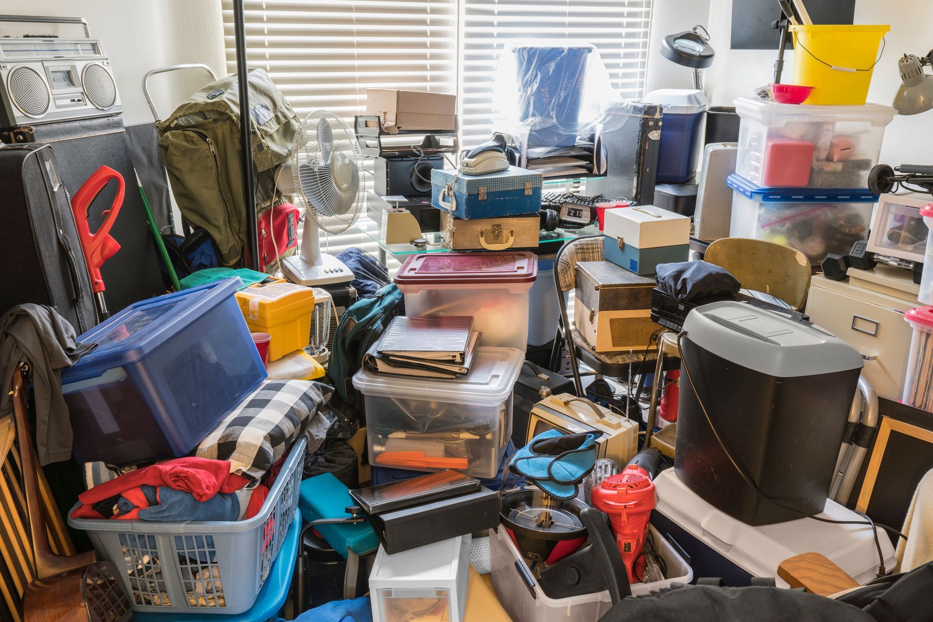 A cluttered room filled with stacked storage bins, boxes, a laundry basket, a printer, and household items.