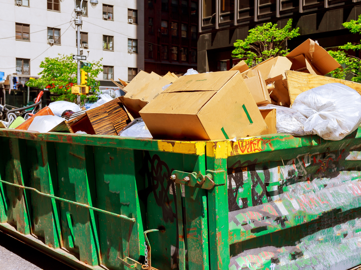 A green dumpster filled with cardboard boxes and plastic bags.