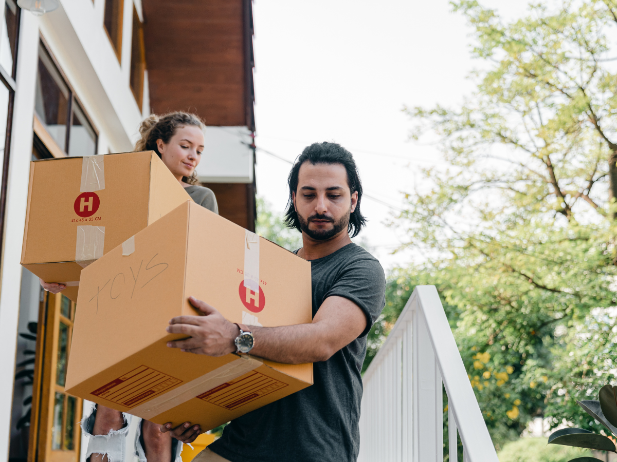 A couple moving boxes out of a house. The man is carrying a large box, and the woman carries a smaller one.