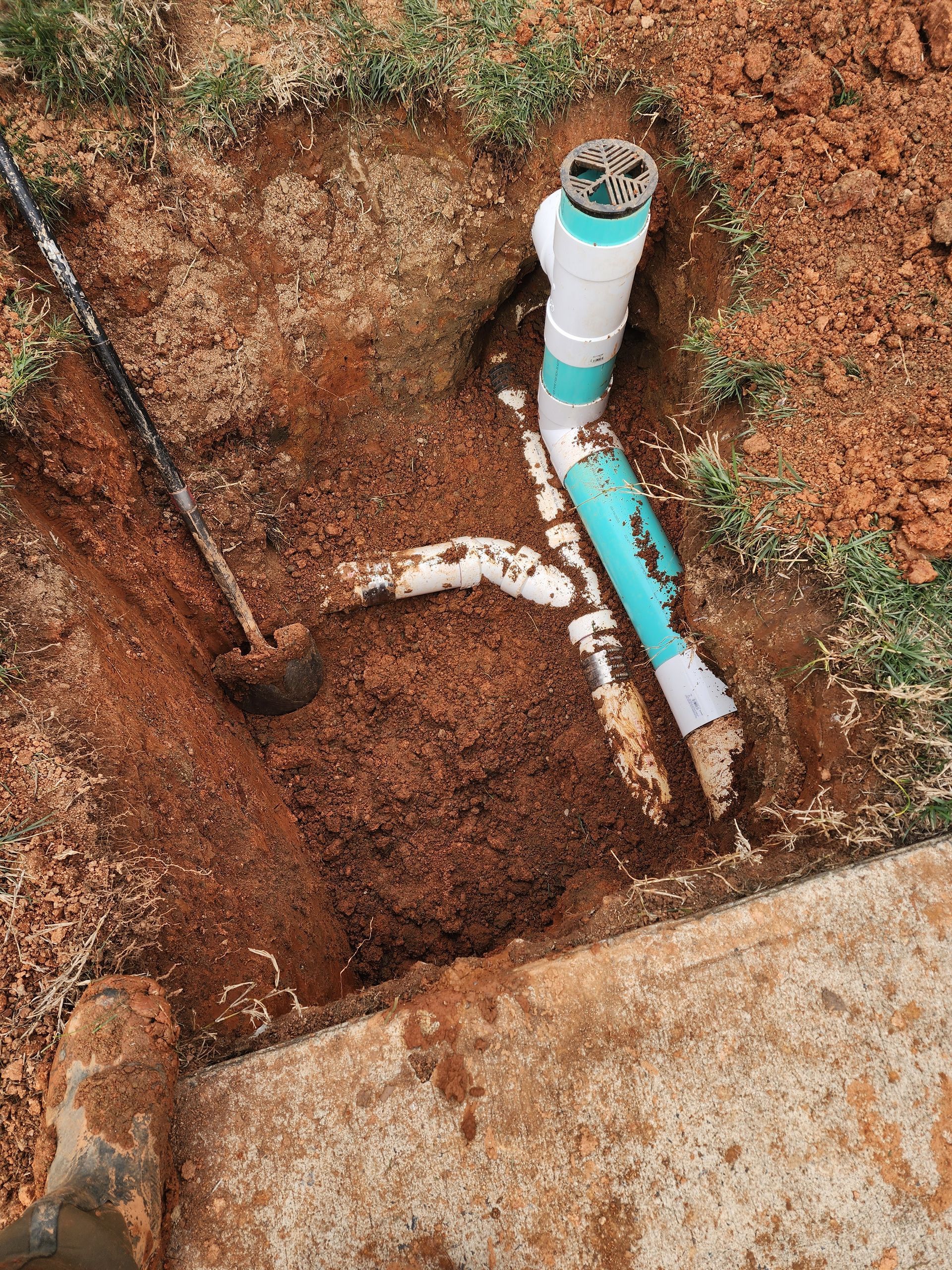 Plumbing pipes exposed in a dirt hole near a concrete edge; shovel nearby.