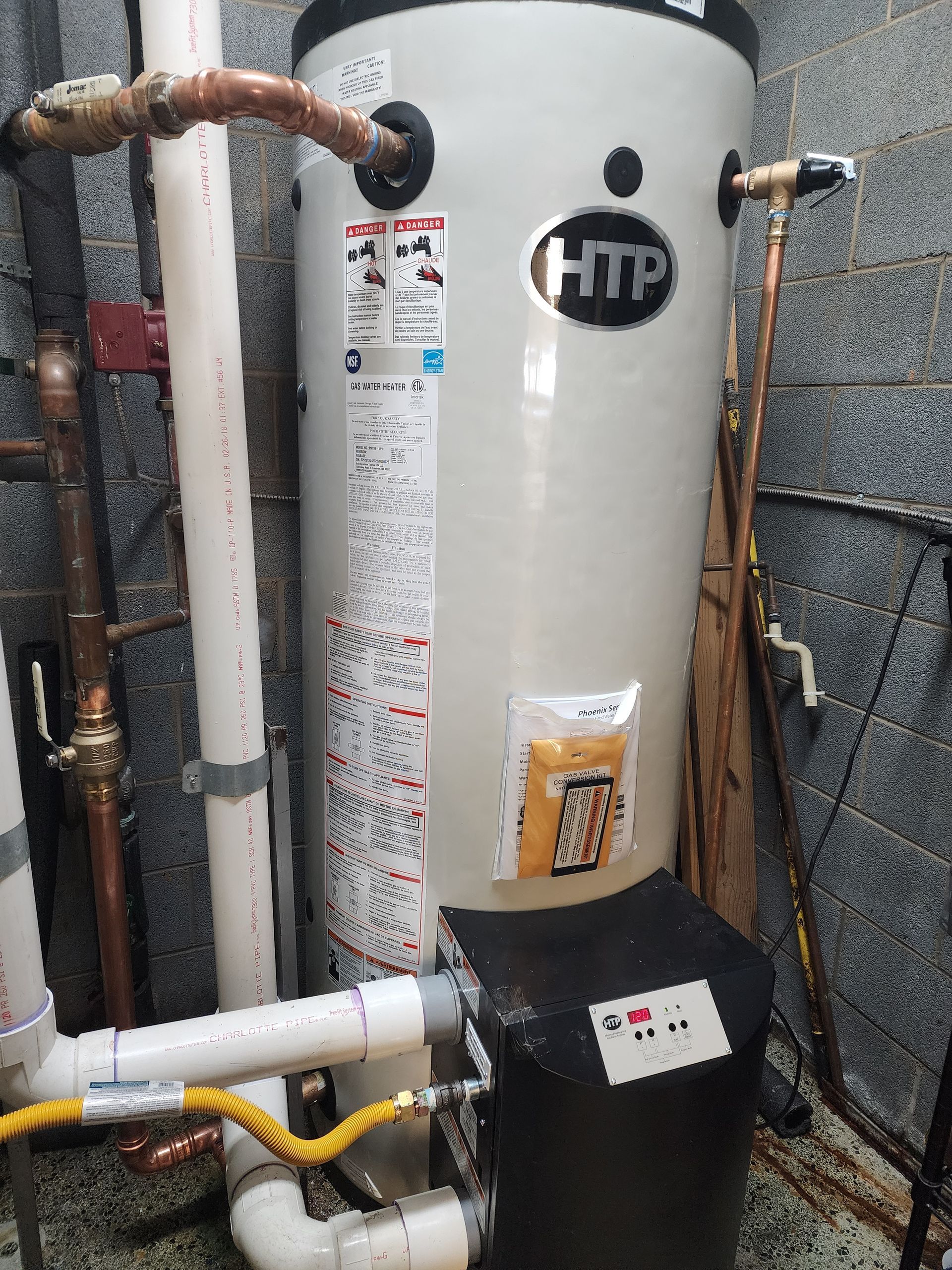 Tall, cylindrical HTP water heater with white and black components, copper pipes, and a PVC pipe in a utility room.