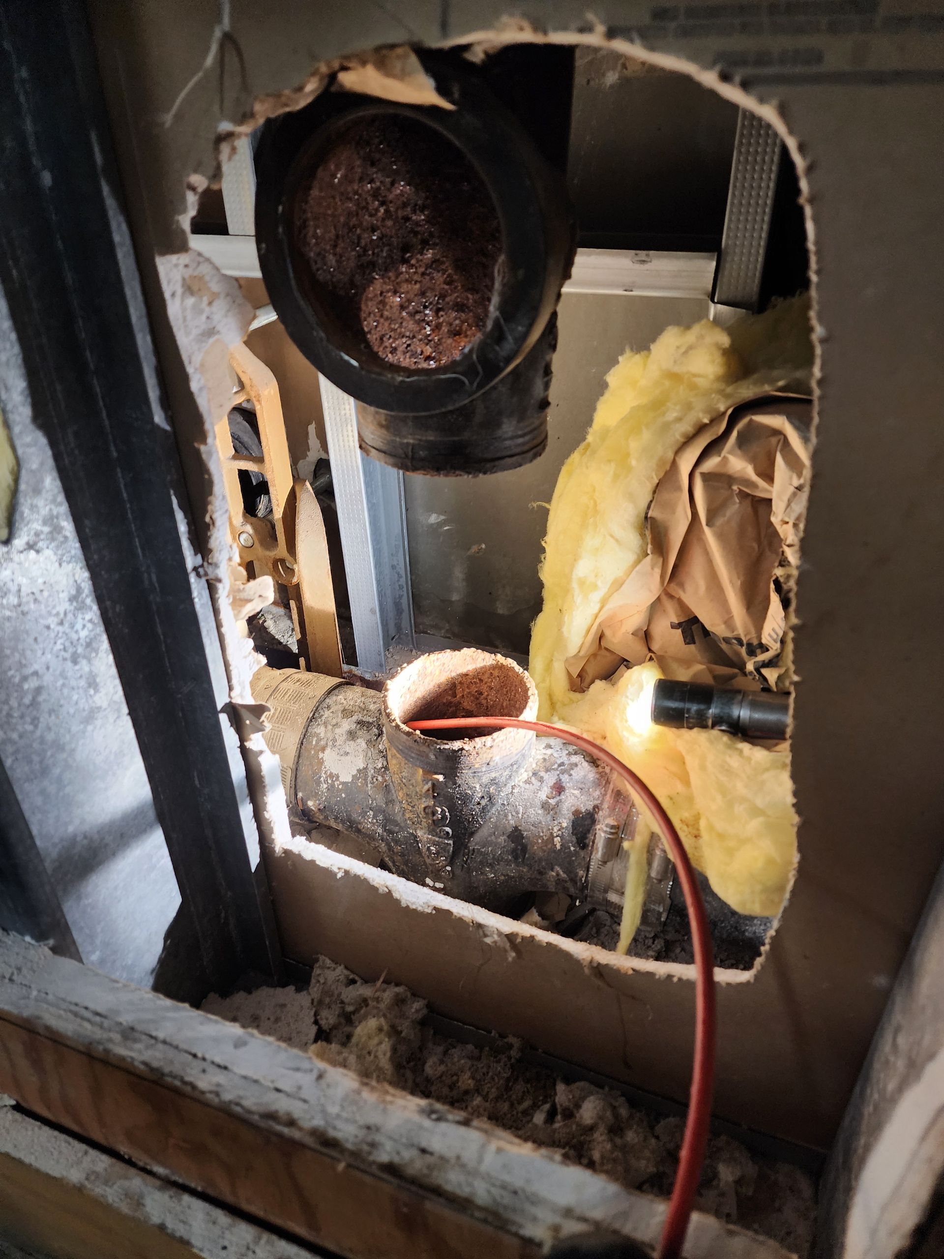 Hole in wall exposing plumbing pipes, insulation, and a flashlight; brown and black tones.