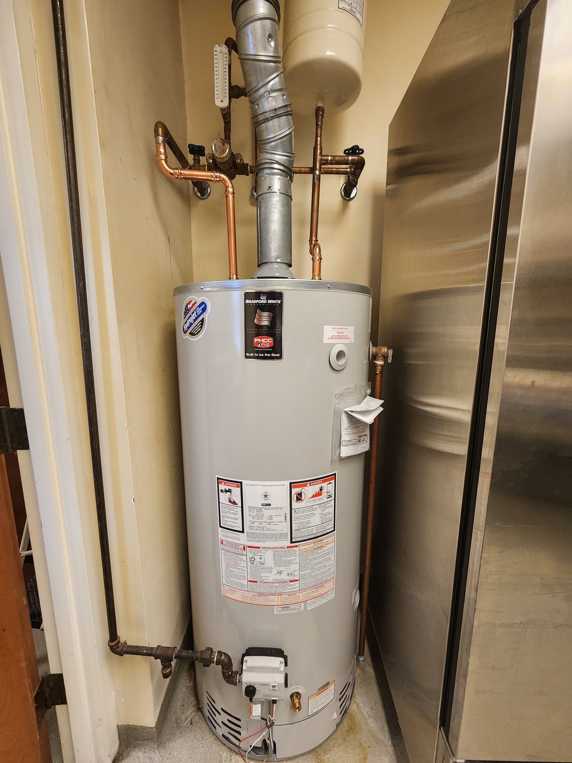 A gas water heater in a utility room with copper pipes and a silver tank.
