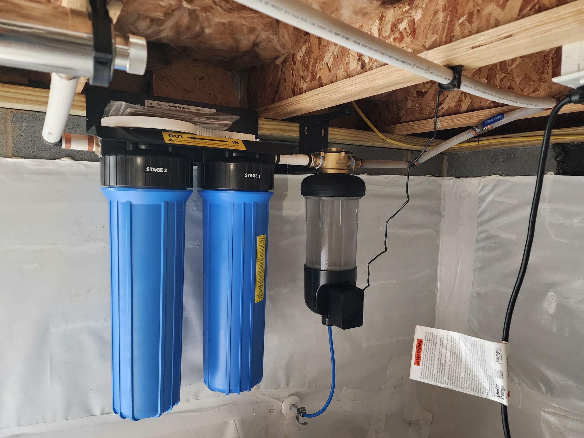 Blue water filter system installed on a basement ceiling. Includes three filter housings, pipes, and wiring.