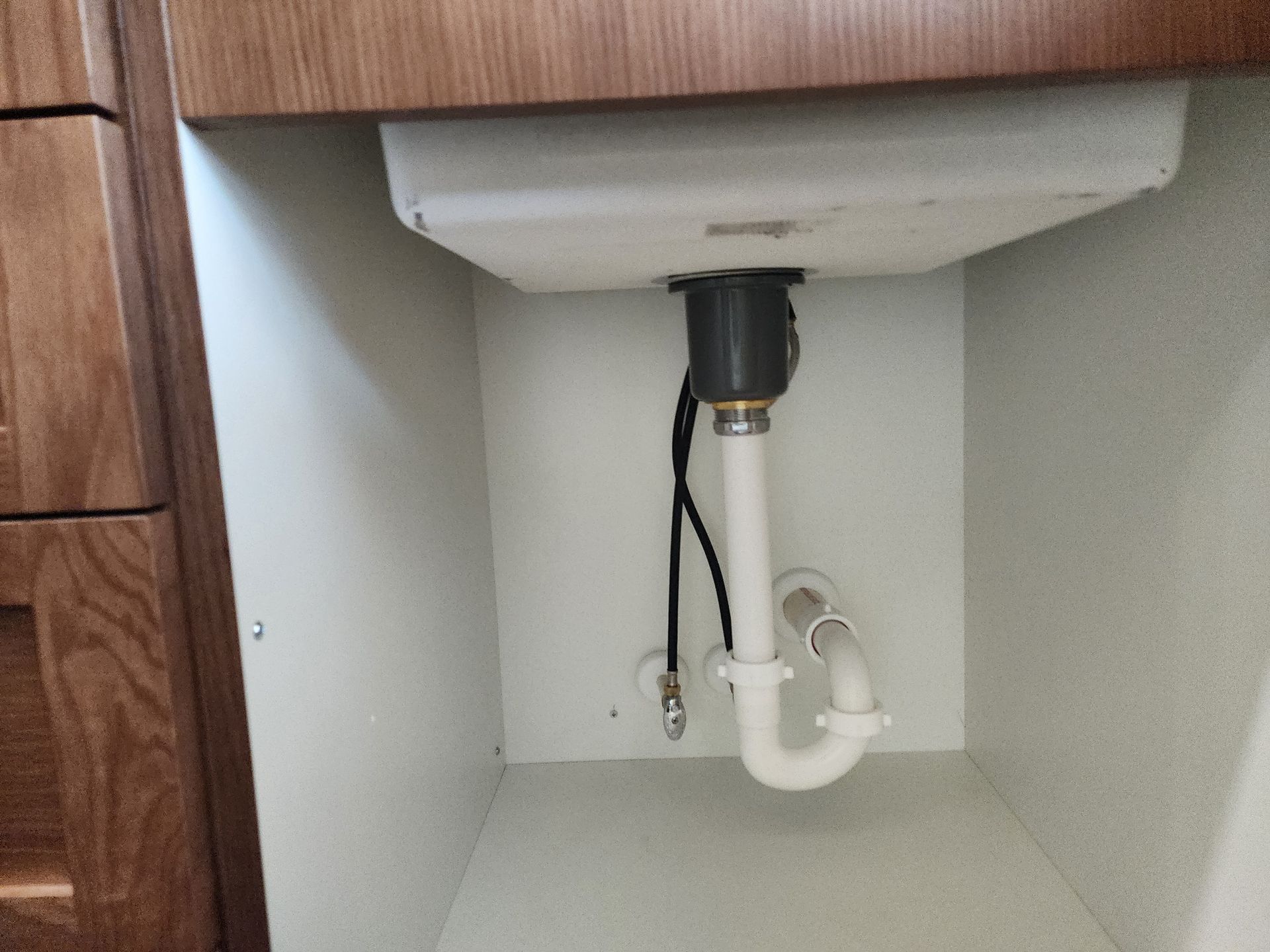 Under-sink plumbing: white pipes, a gray drain, and a black hose inside a cabinet with a wooden countertop.