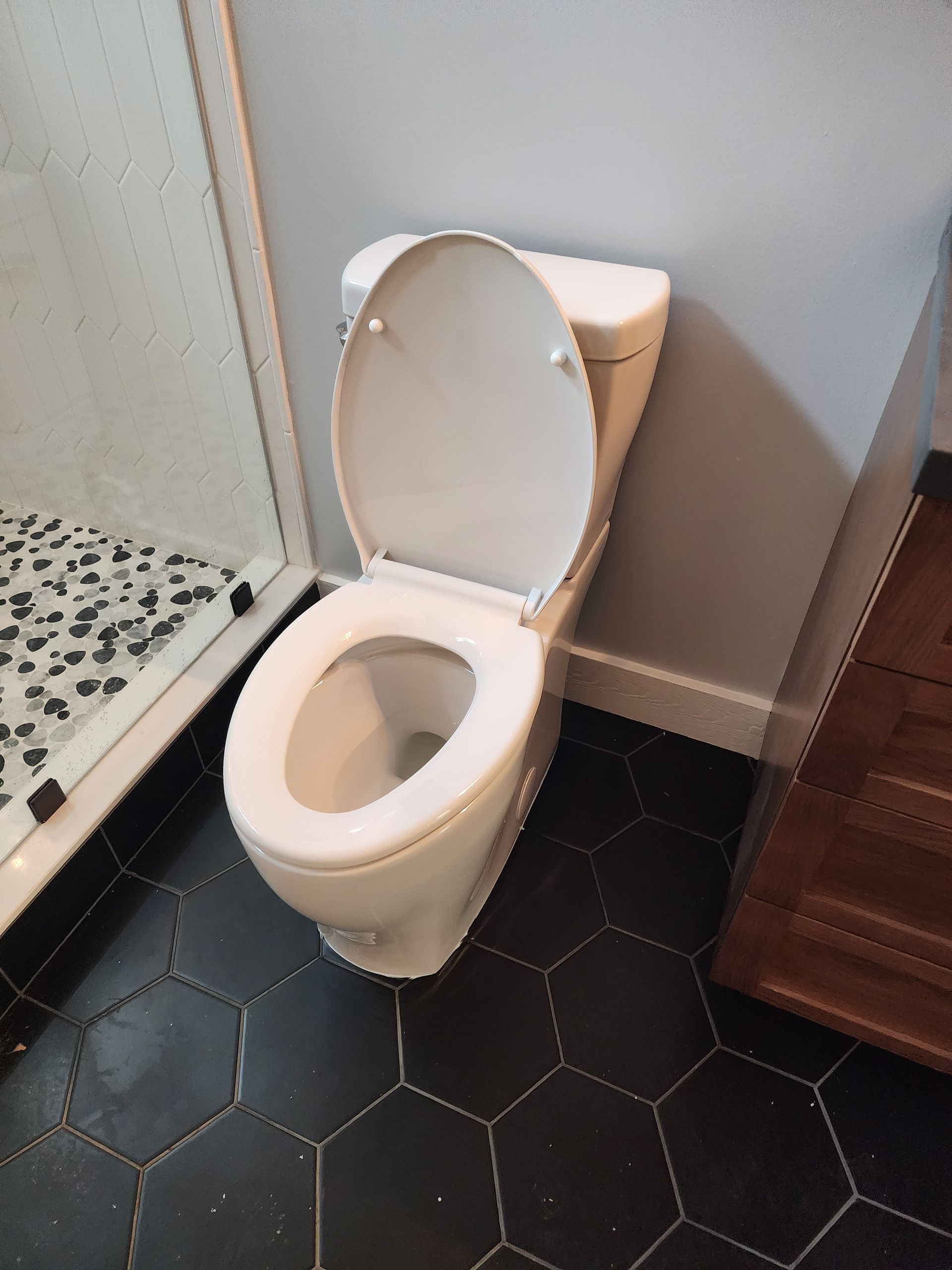 White toilet with open lid in a bathroom with black hexagon tiles, next to a shower and wooden cabinet.