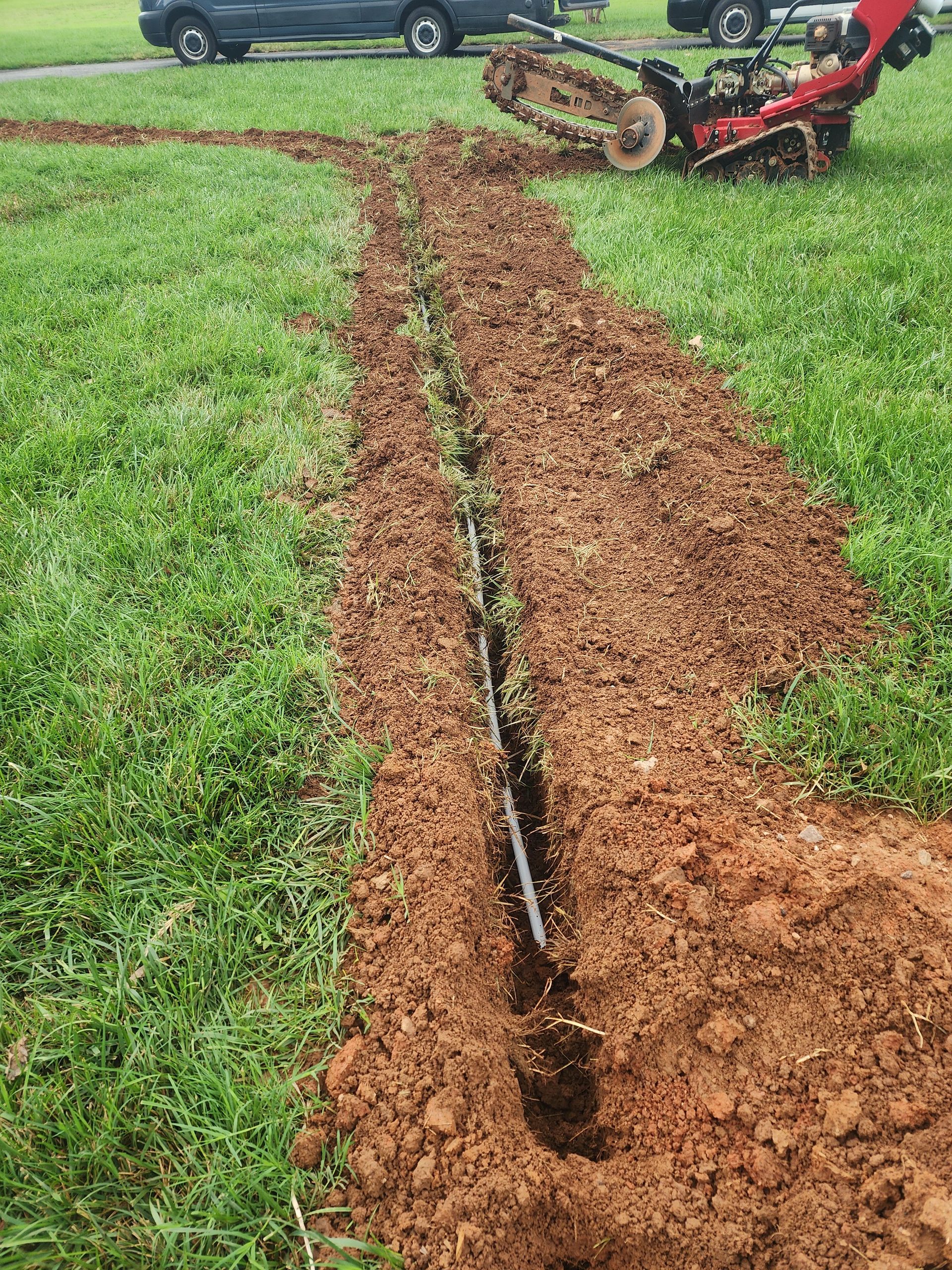 A trencher digging a trench in a grassy field, with a black pipe visible in the freshly dug soil.