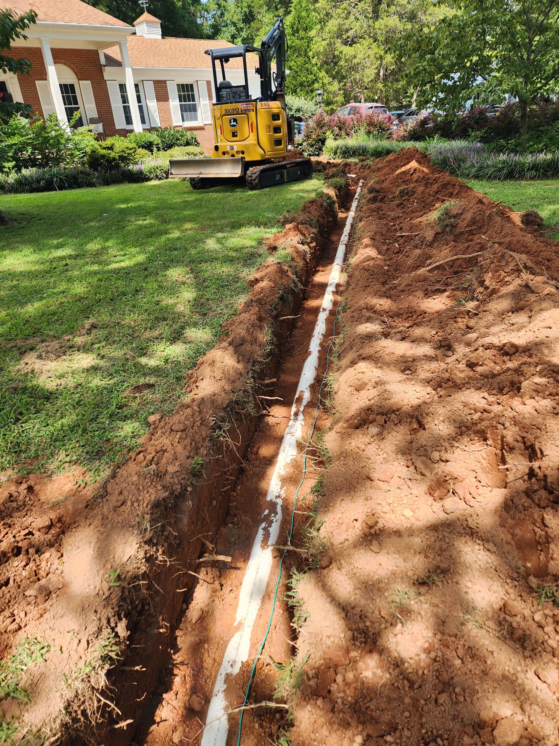 Trench dug in a yard with white pipe visible, mini excavator in background near a house.