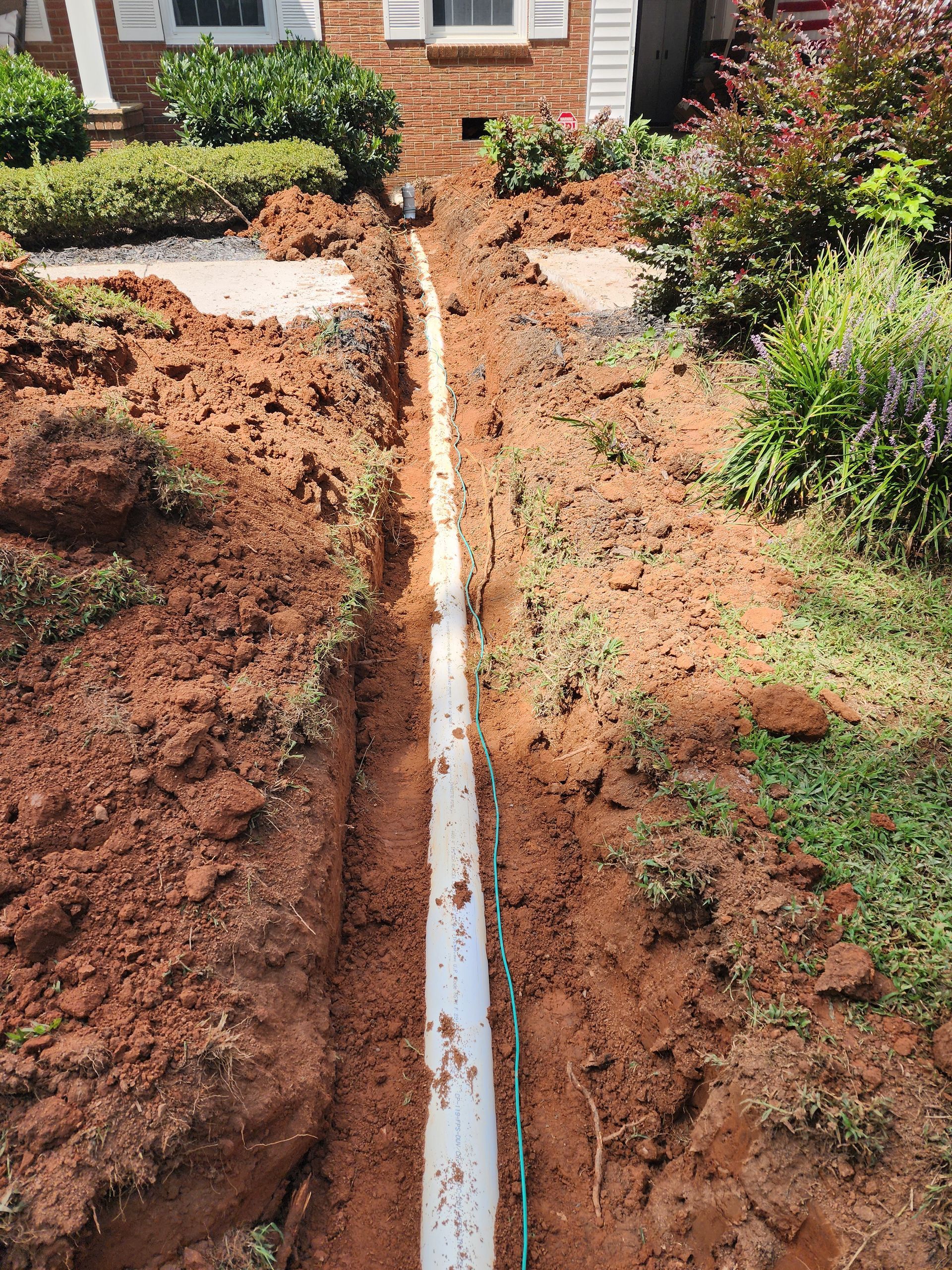 Trench dug in red soil, with white pipe laid, in front of a house.