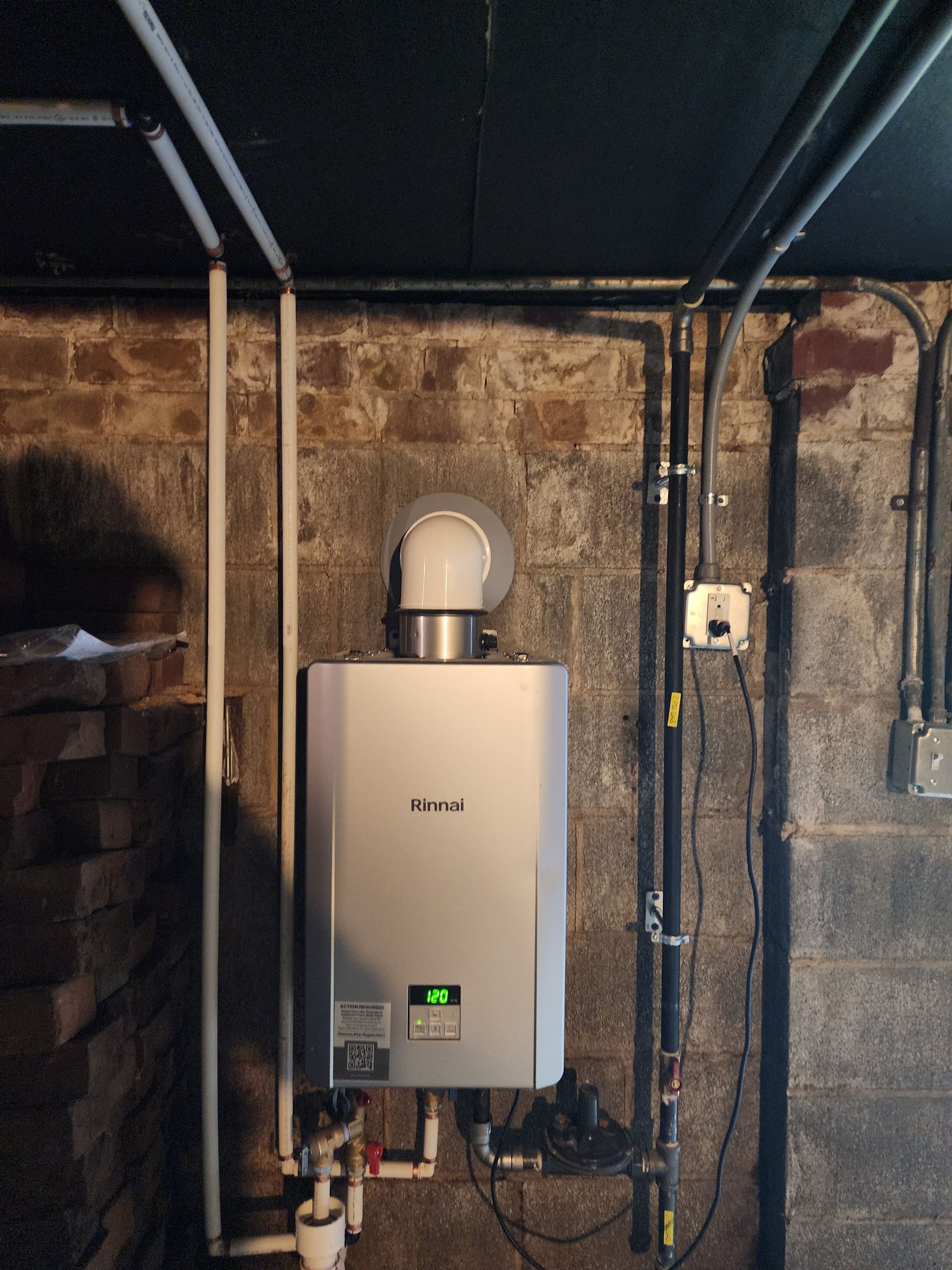 Wall-mounted silver tankless water heater with vent pipe, installed in a basement with brick walls.