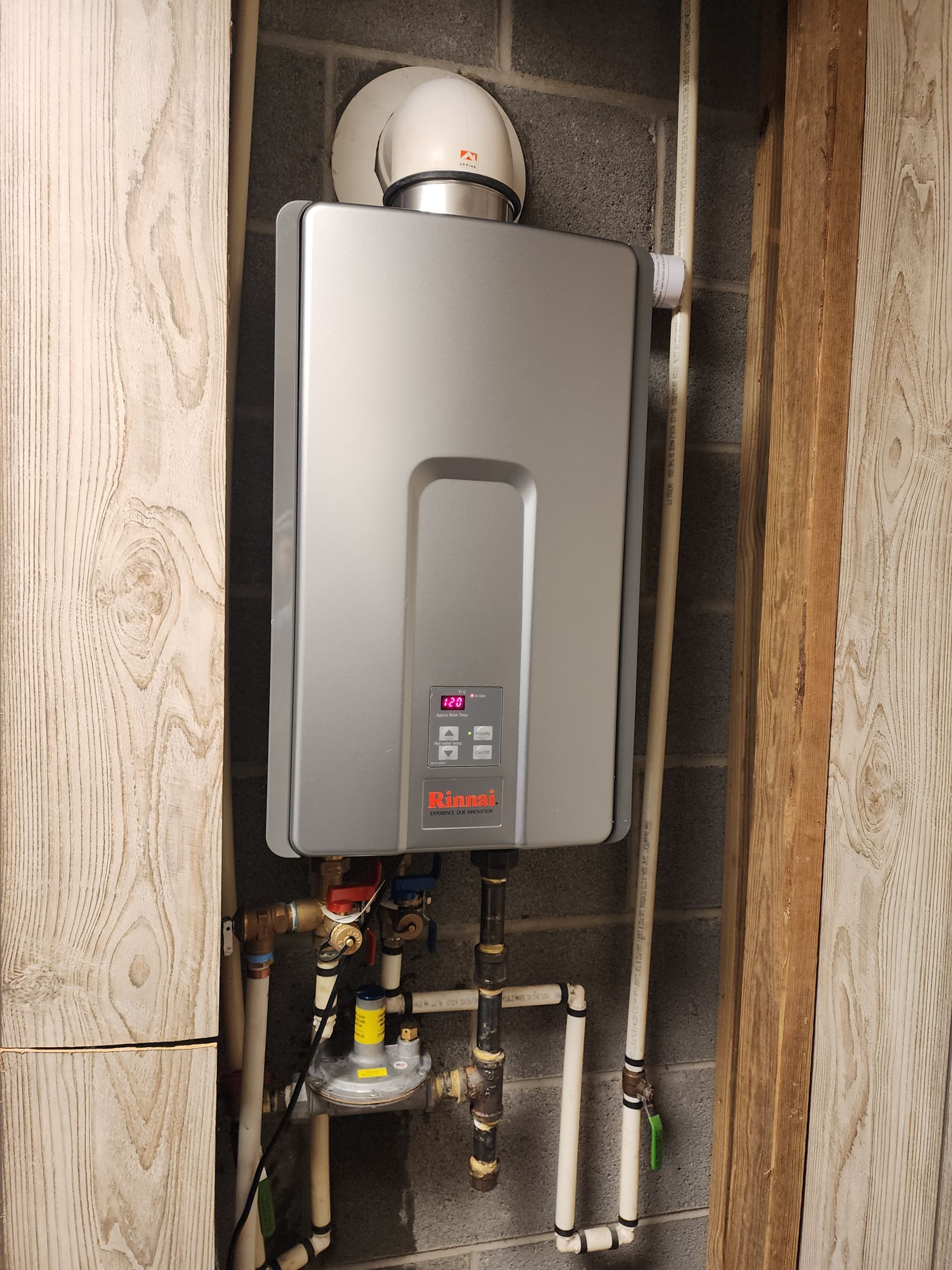 Silver tankless water heater mounted on a gray brick wall, between wooden beams.