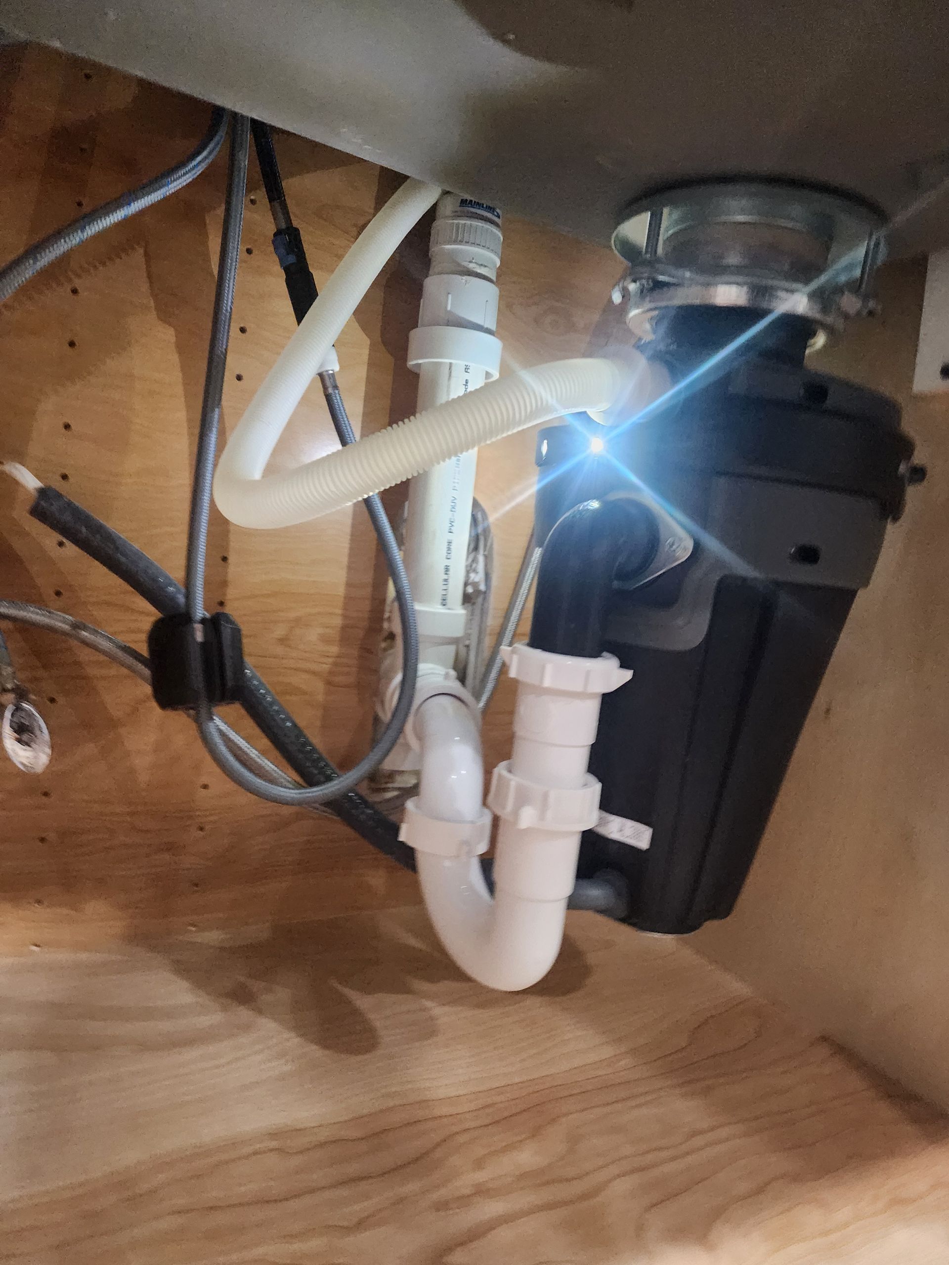Under-sink plumbing with garbage disposal, white pipes, and wooden cabinet interior.