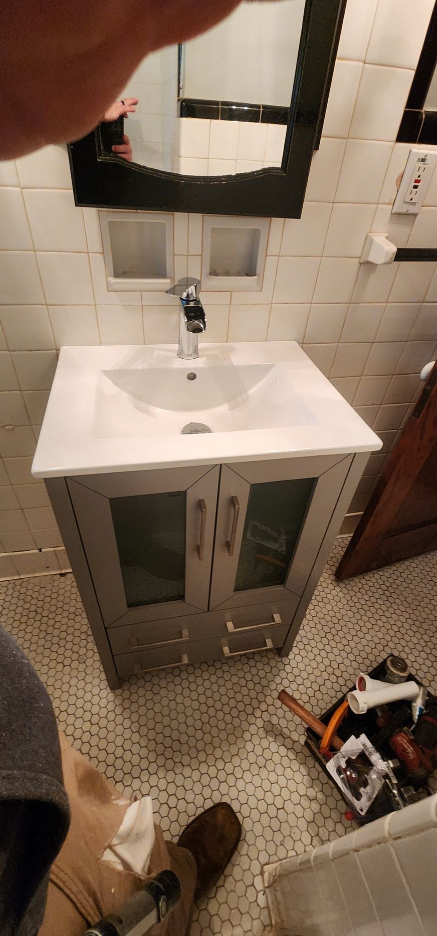 Bathroom vanity with gray cabinet, sink, faucet, mirror, and tools on floor. White tile and hexagon floor.