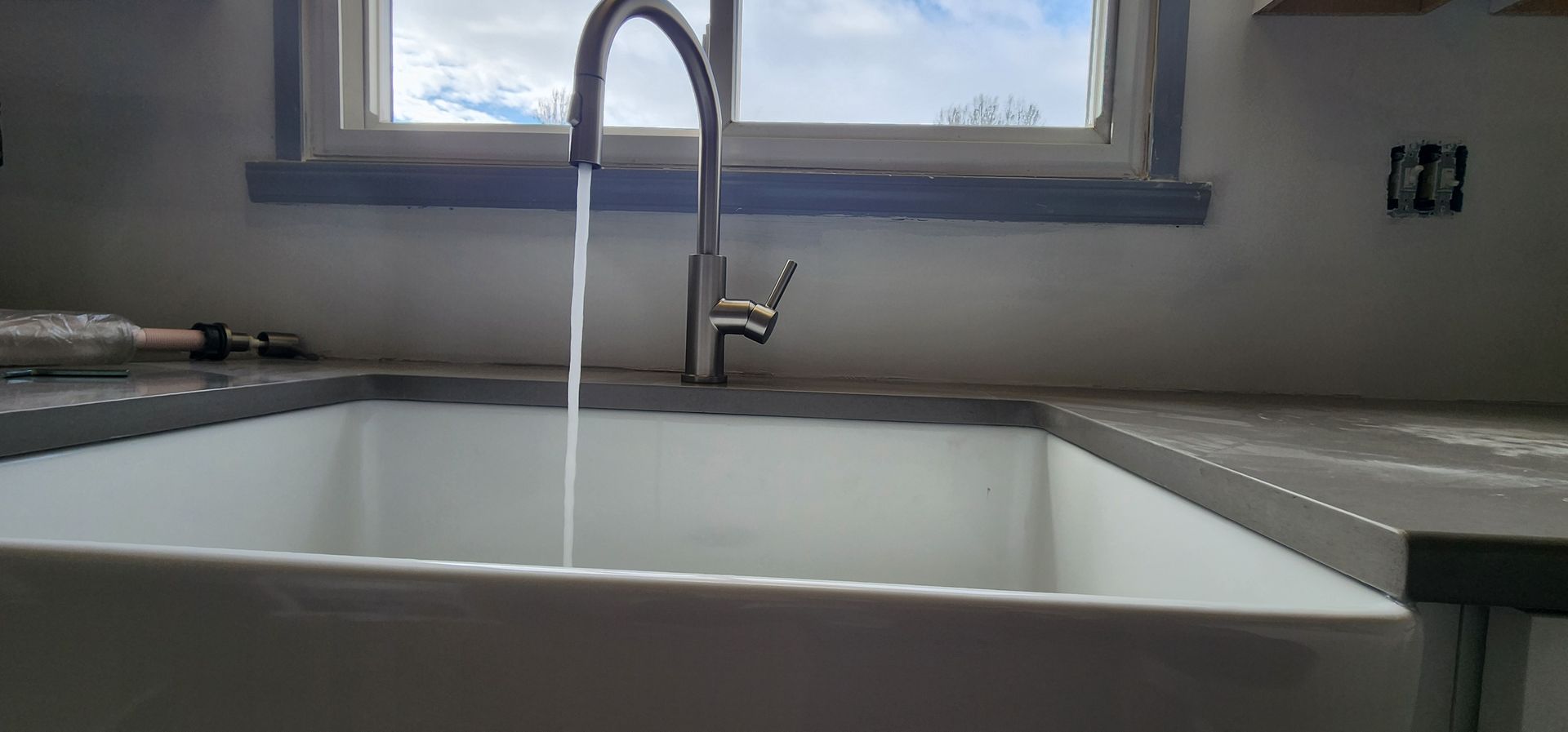 Water flows from a kitchen faucet into a white farmhouse sink.  A window is behind the faucet.