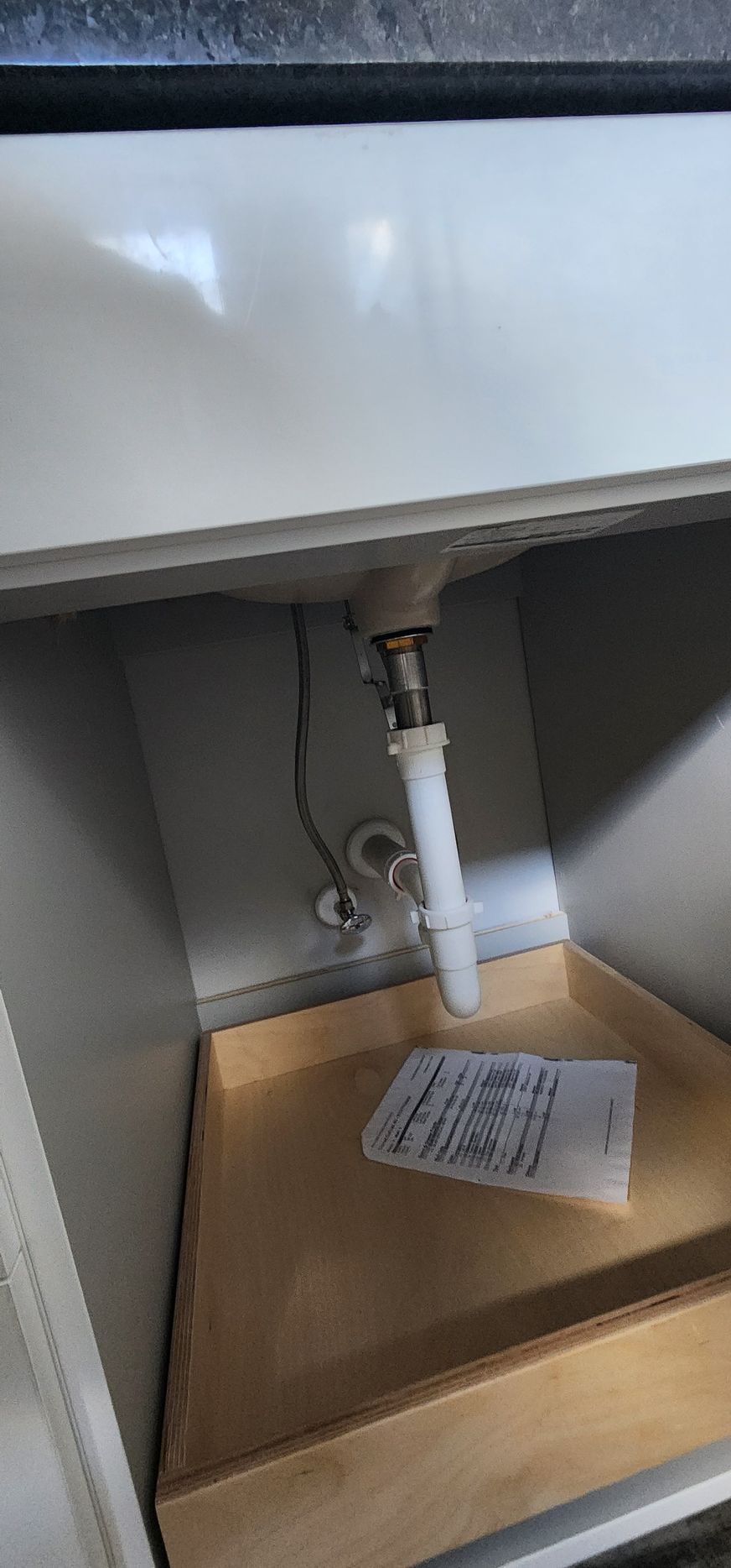 Inside a white bathroom sink cabinet; drain pipes, and a partially pulled-out wooden drawer are visible.