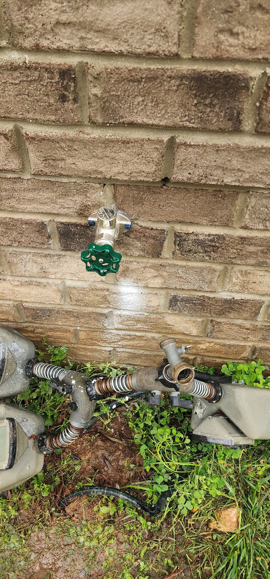 Outdoor spigot with a green handle, brick wall background, and hose connected to a container on the ground.