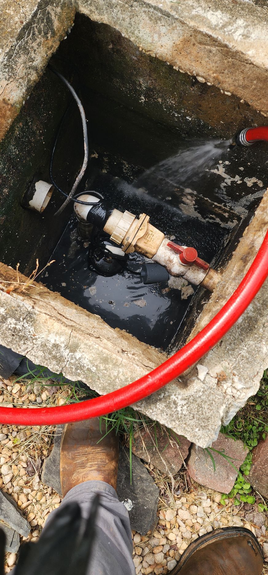 Red hose and valve spraying water into a concrete pit. Dark liquid inside. Person's boots visible.