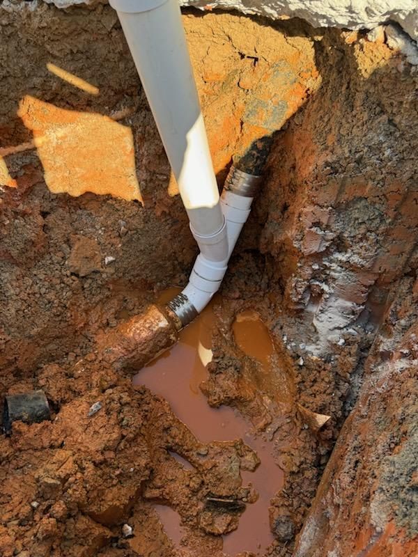 Gray pipes in a muddy trench. Water pools around a Y-shaped pipe fitting.
