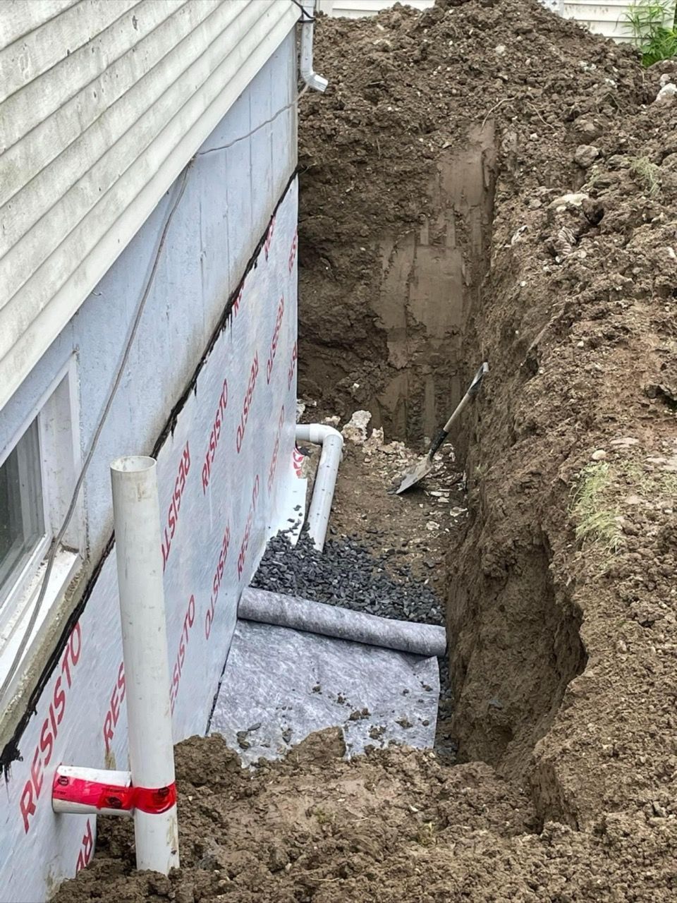 Un mur de fondation est en cours de travaux. Une tranchée a été creusée à côté du mur, les éléments de drainage sont visibles et un nouveau pare-vapeur a été installé.