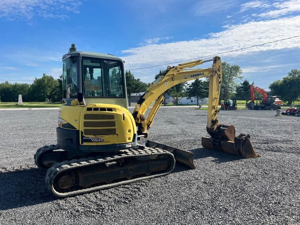 Une mini-pelle jaune est garée à l'extérieur, sur du gravier sous un ciel bleu.