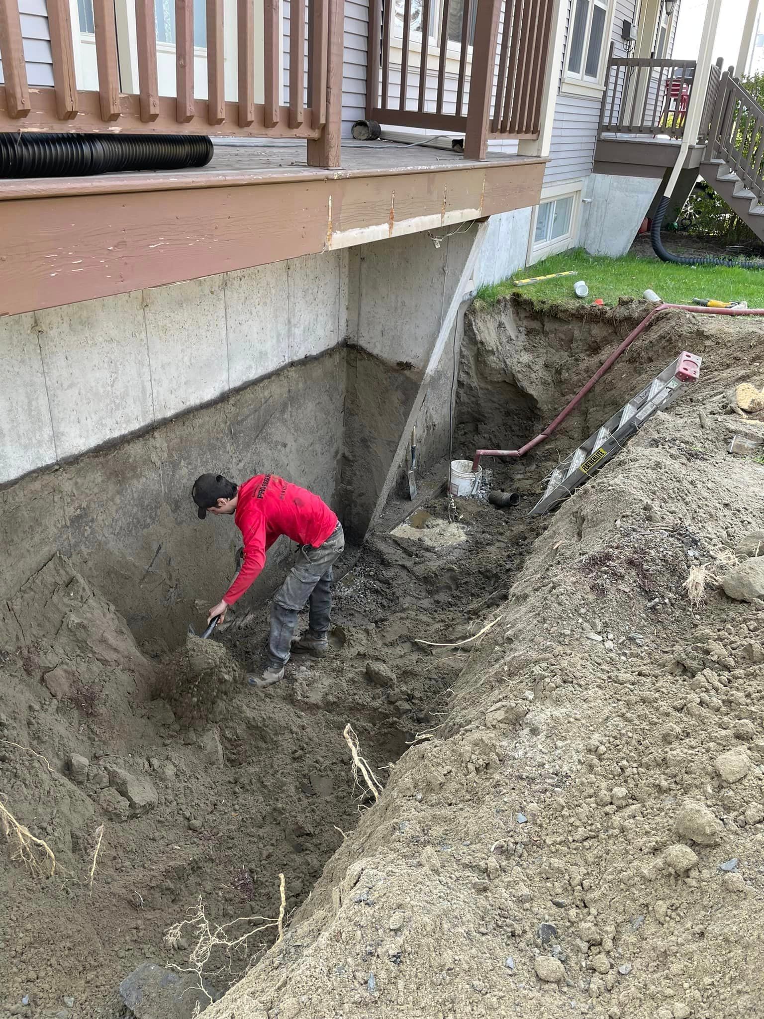 Une personne en chemise rouge creuse la terre près d'une fondation en béton sous une terrasse en bois.