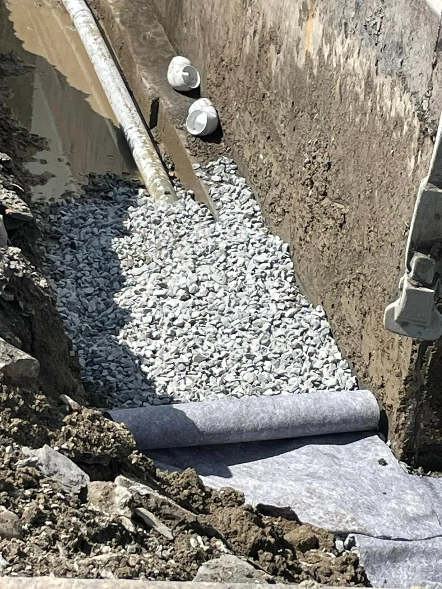 Tranchée remplie de gravier, tapissée de tissu, à côté d'un tuyau et de terre, probablement à proximité d'un chantier.