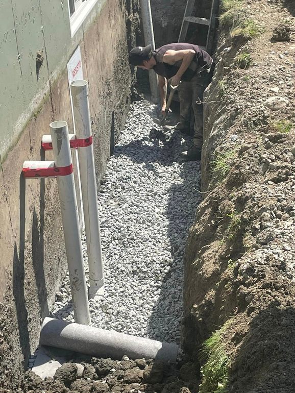Un homme pelle dans une étroite tranchée remplie de gravier près d'un bâtiment. On aperçoit des tuyaux en PVC et une échelle.