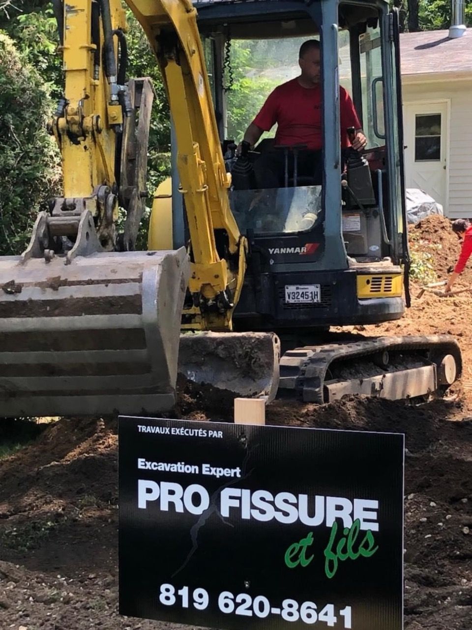 Un homme manœuvre une pelleteuse jaune. Panneau « Pro Fissure et fils » devant. Extérieur.
