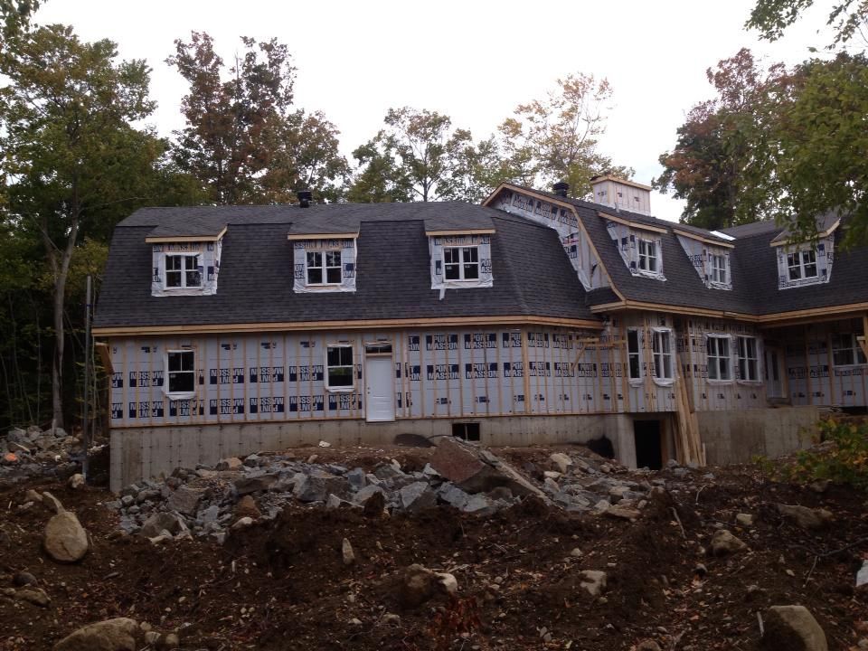 Maison en construction avec toit gris, lucarnes blanches et charpente en bois. Terre brune au premier plan.