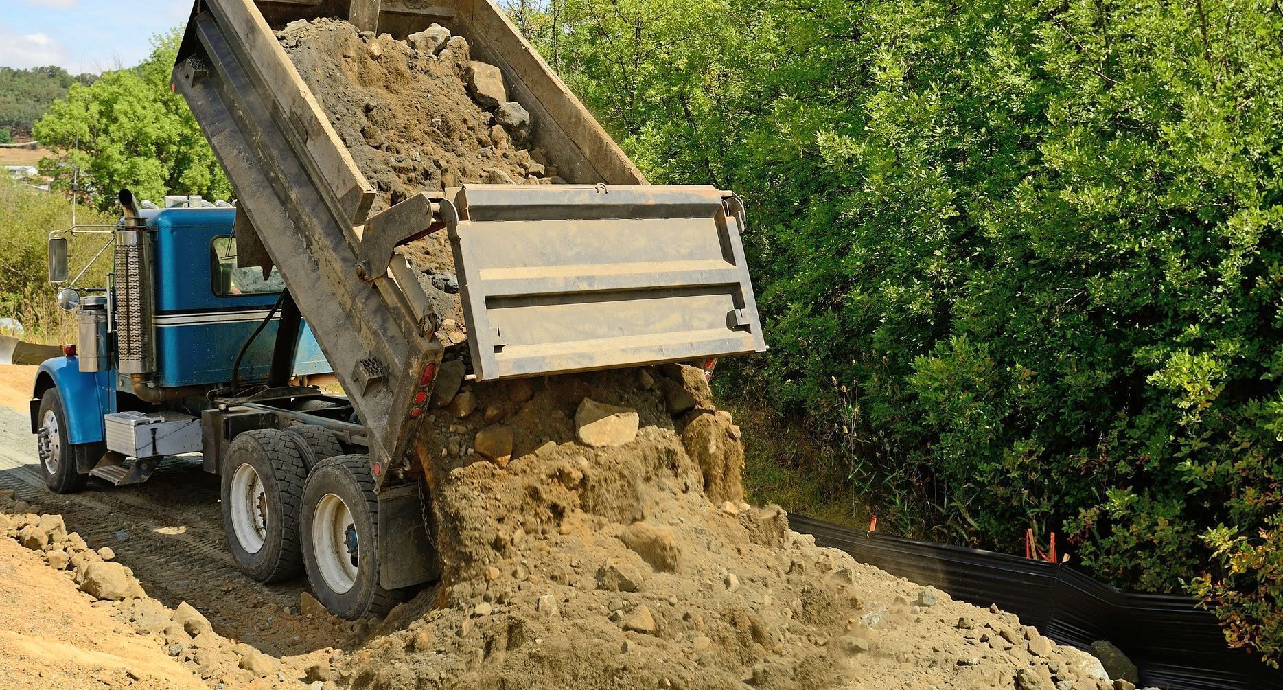 Un camion-benne décharge de la terre en bord de route, près d'arbres verts.