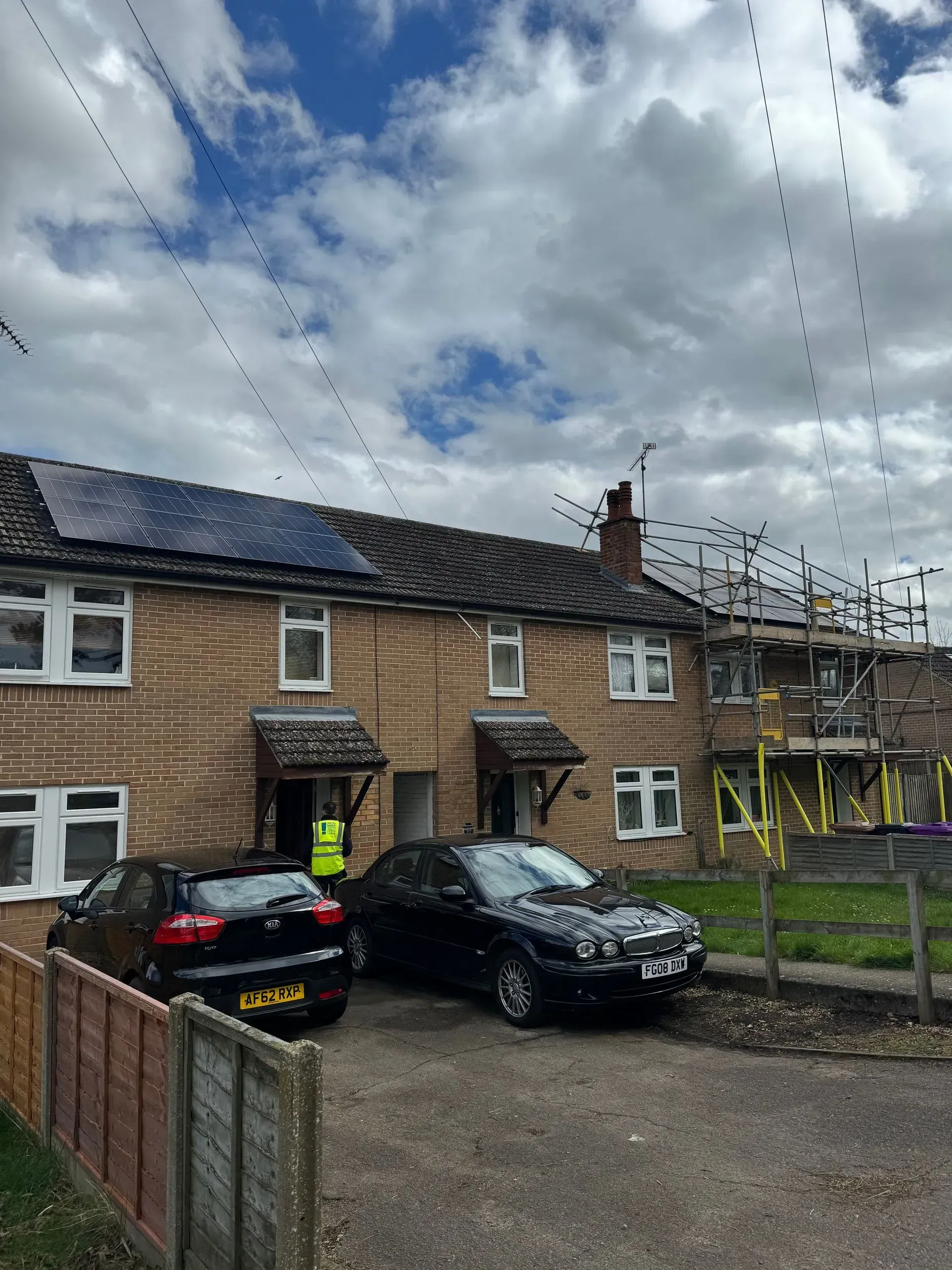 Row of brick houses with solar panels, scaffolding on right, cars parked in front, utility lines above.