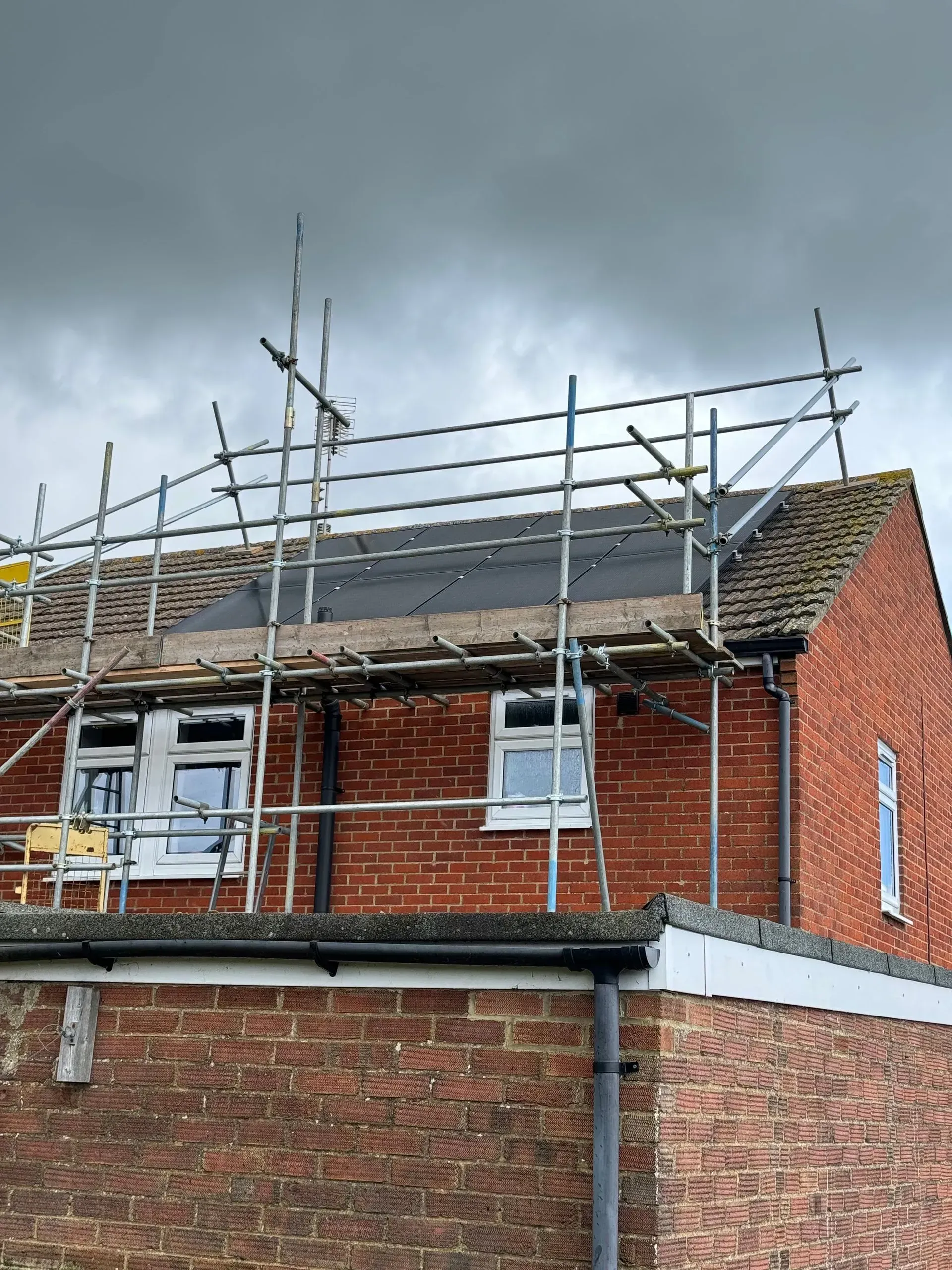 Scaffolding on a brick house roof under a cloudy sky. Roofing work in progress.