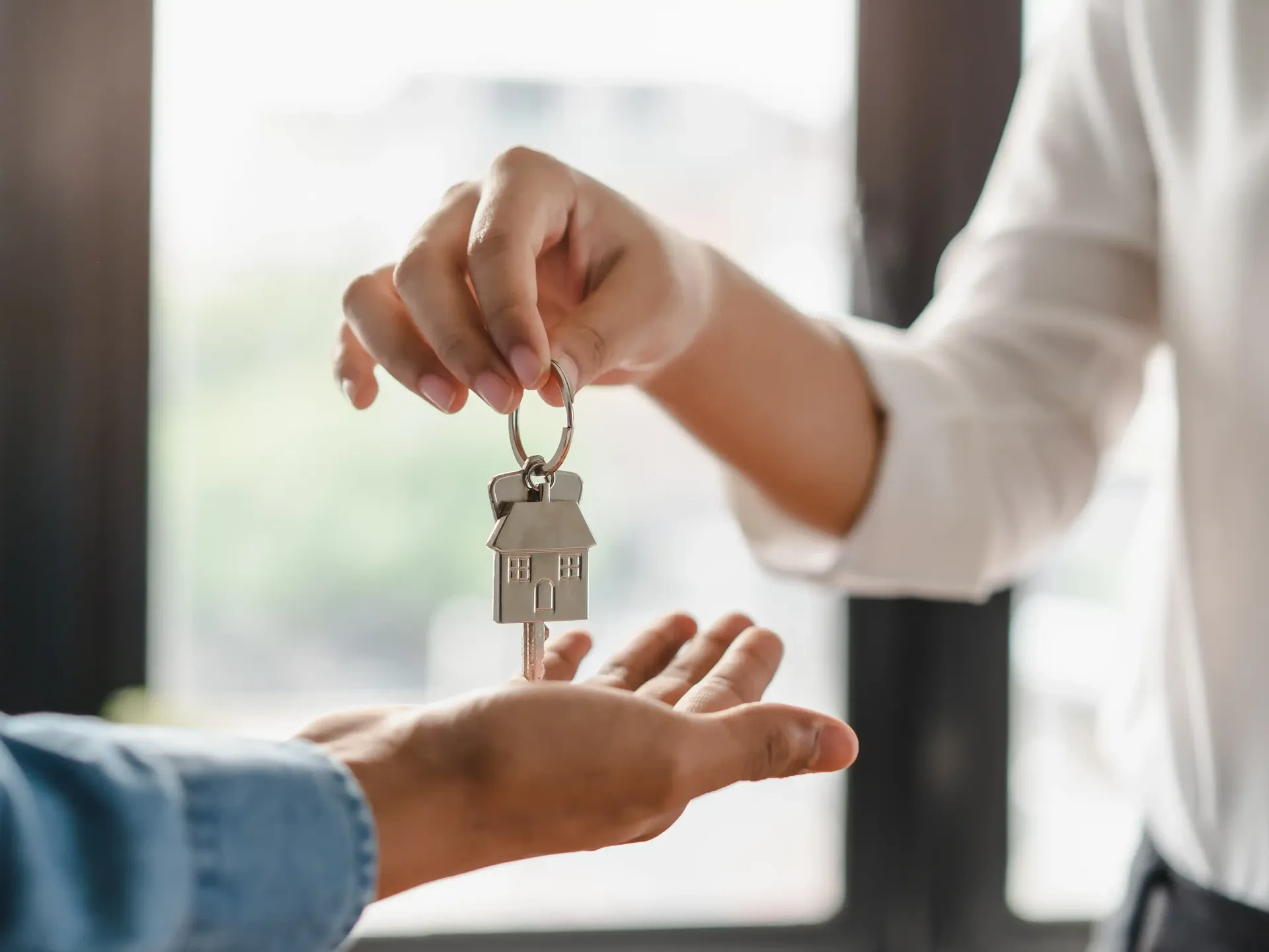 Person handing keys with house-shaped keychain to another person, near a window.