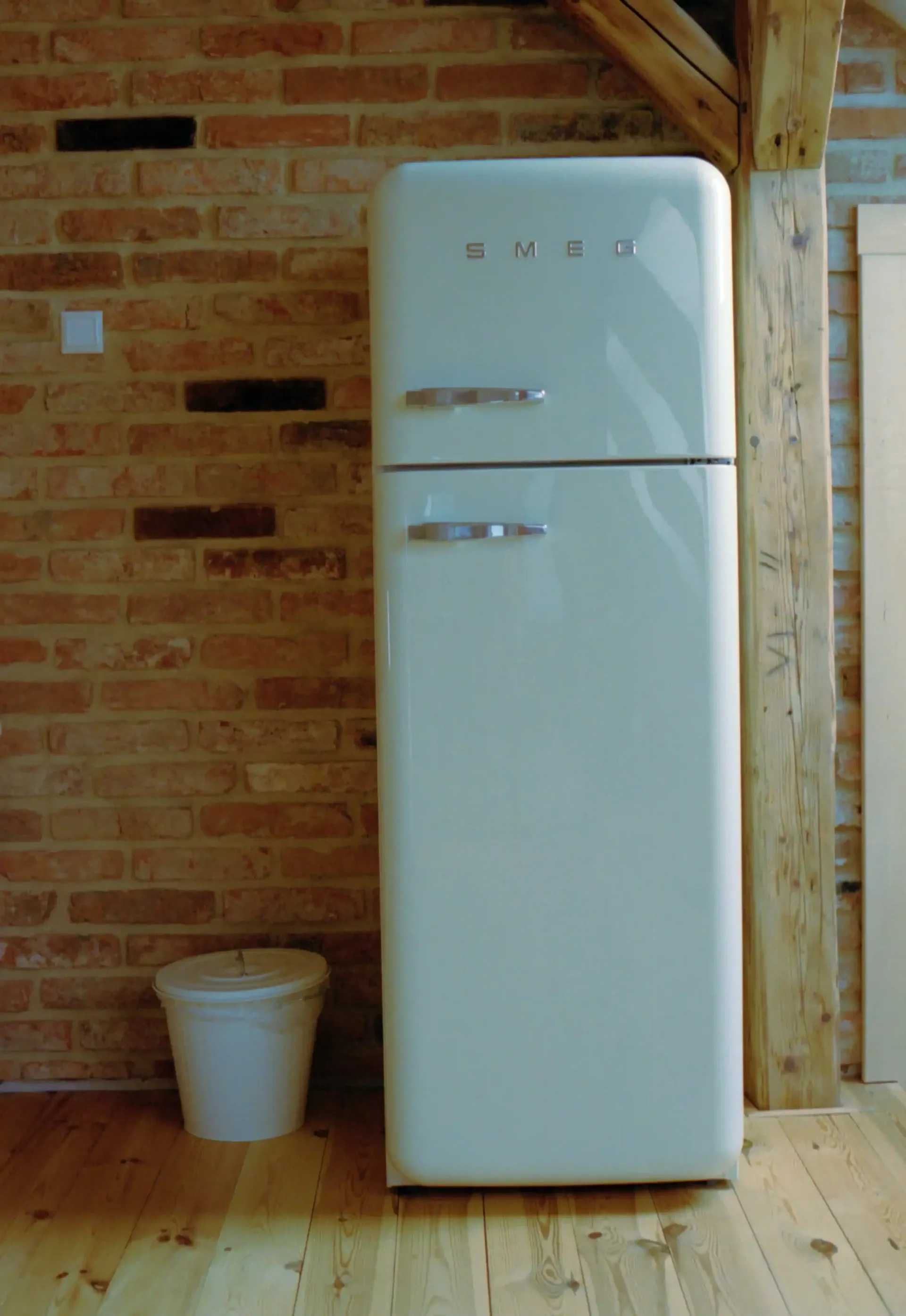 Cream-colored retro refrigerator stands against a brick wall. A small bucket sits on the wooden floor.