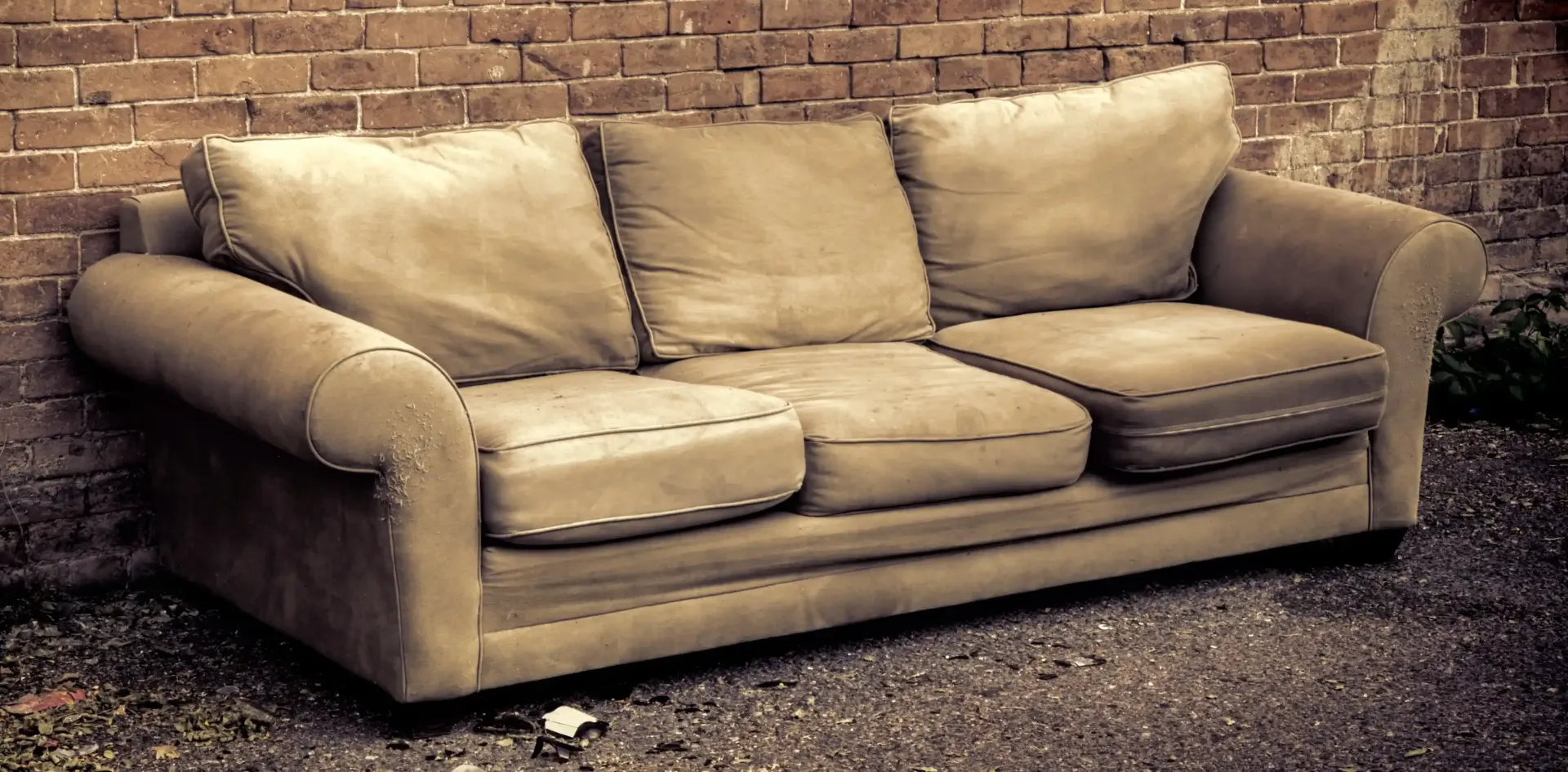 An old, beige couch sitting against a brick wall.