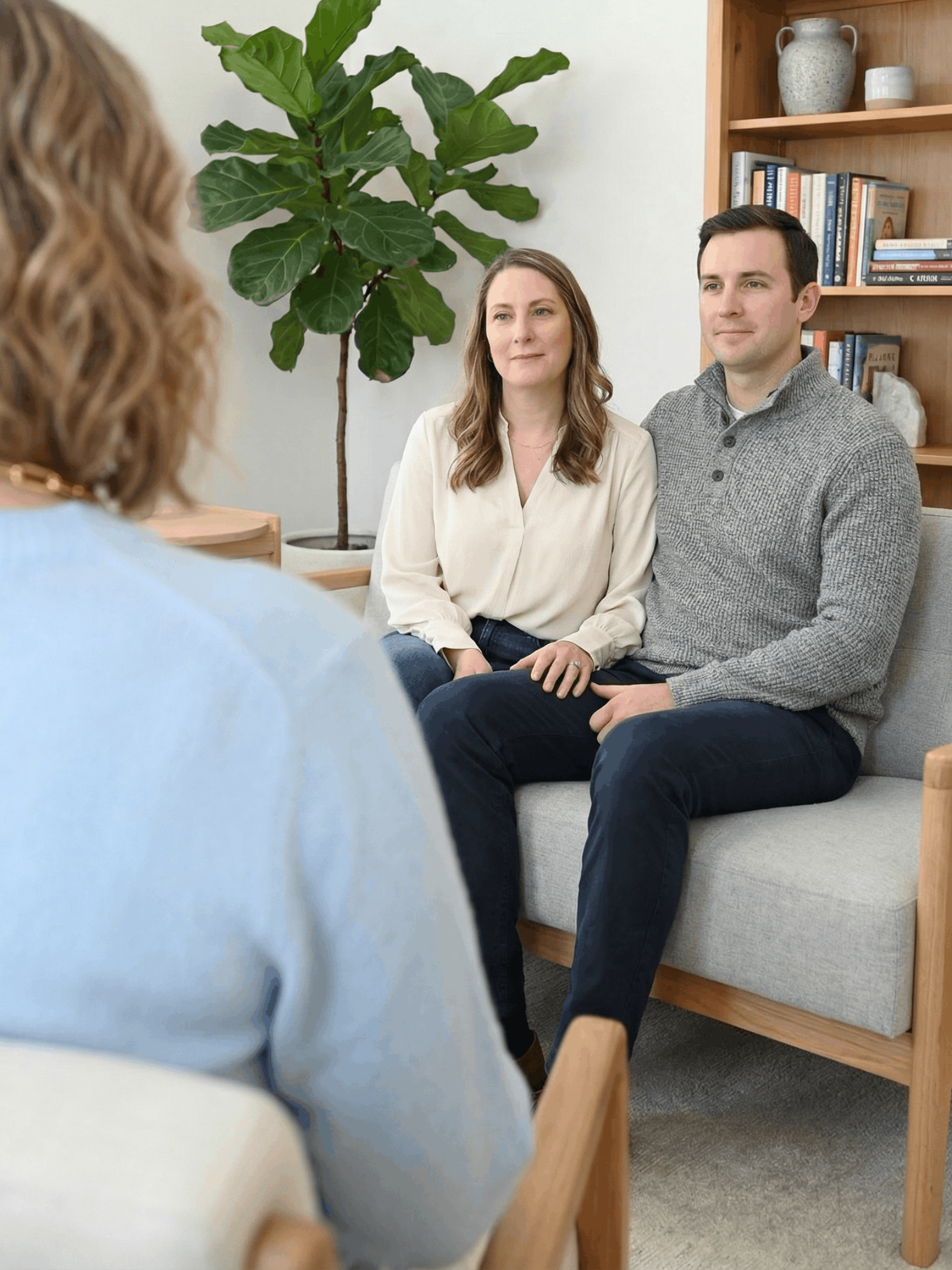 Man seated on a couch with hand on forehead, talking to a person, indoors.