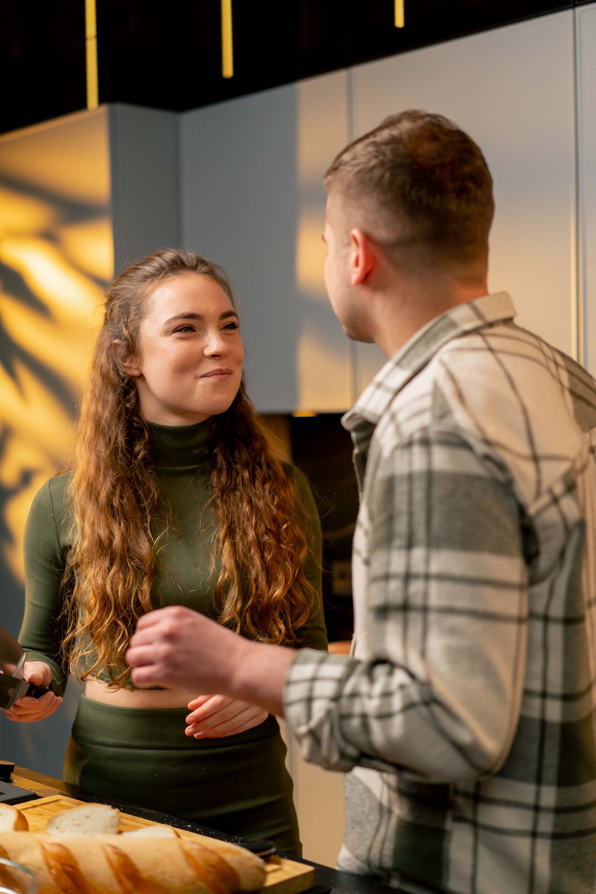 Woman in green top smiles at man in plaid shirt in a kitchen with baguette on a counter.