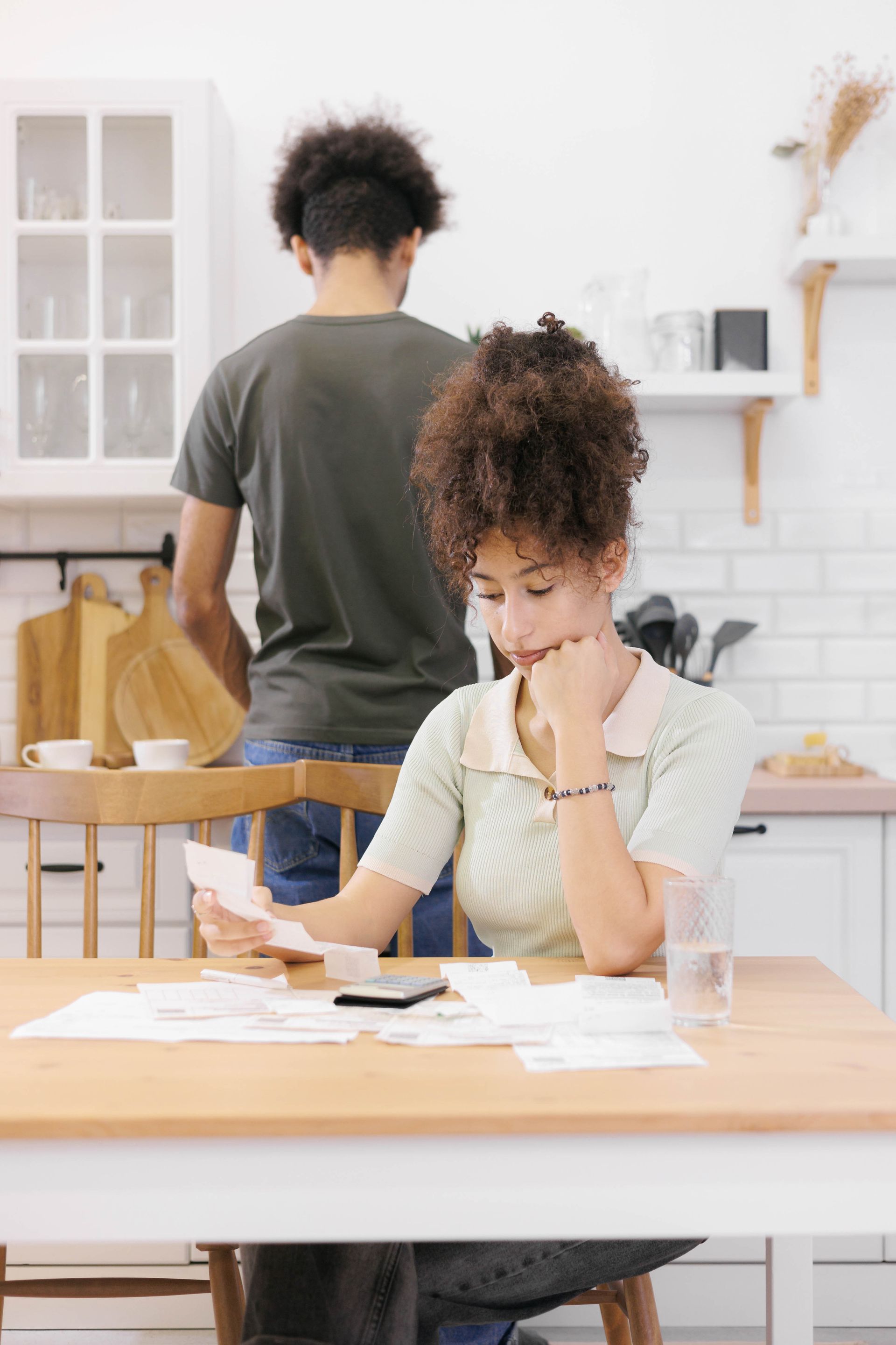 Woman at table reviews bills; man in background, kitchen setting.