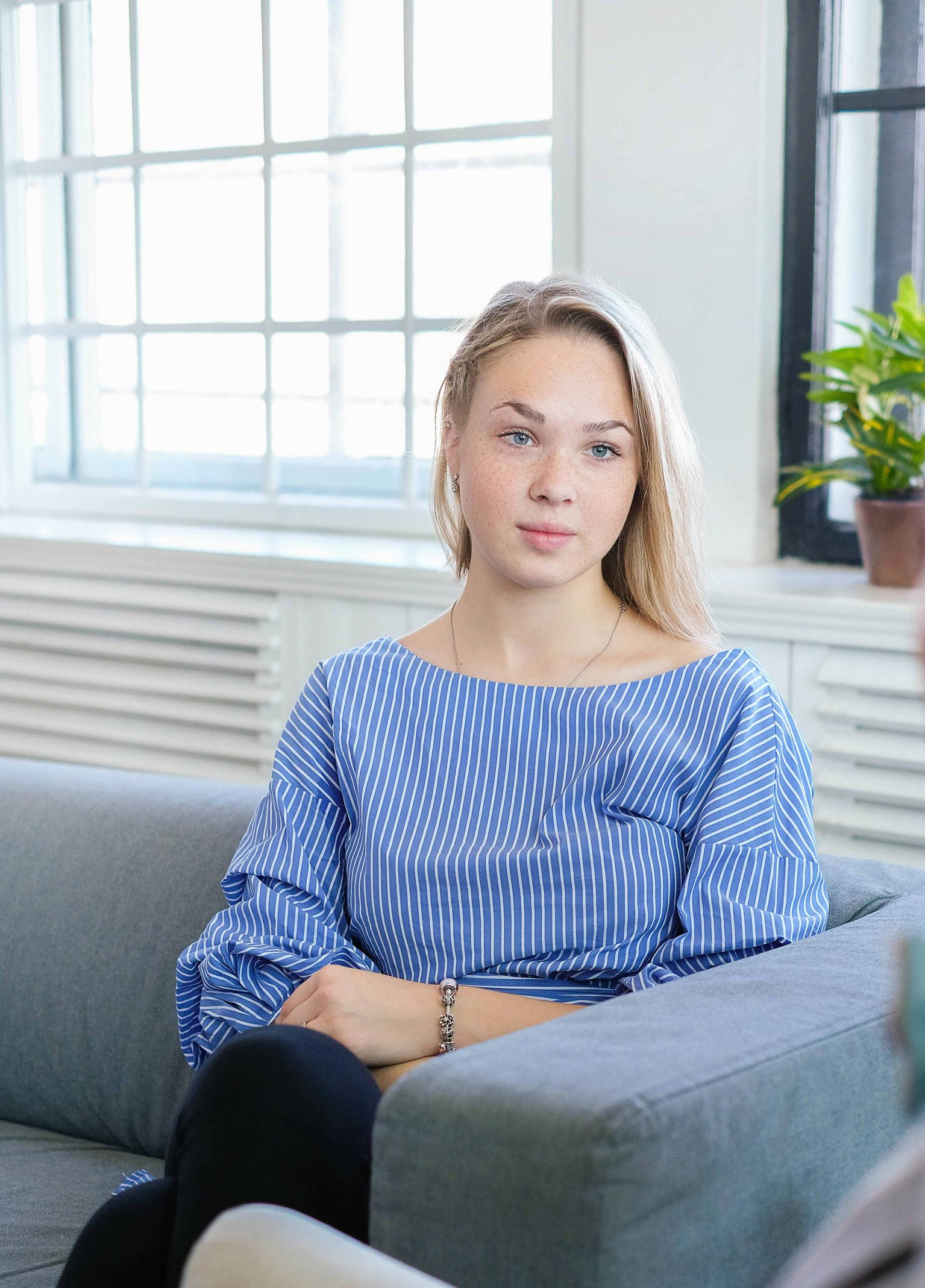 Woman in blue top sits on a gray couch, looking towards right; bright room.