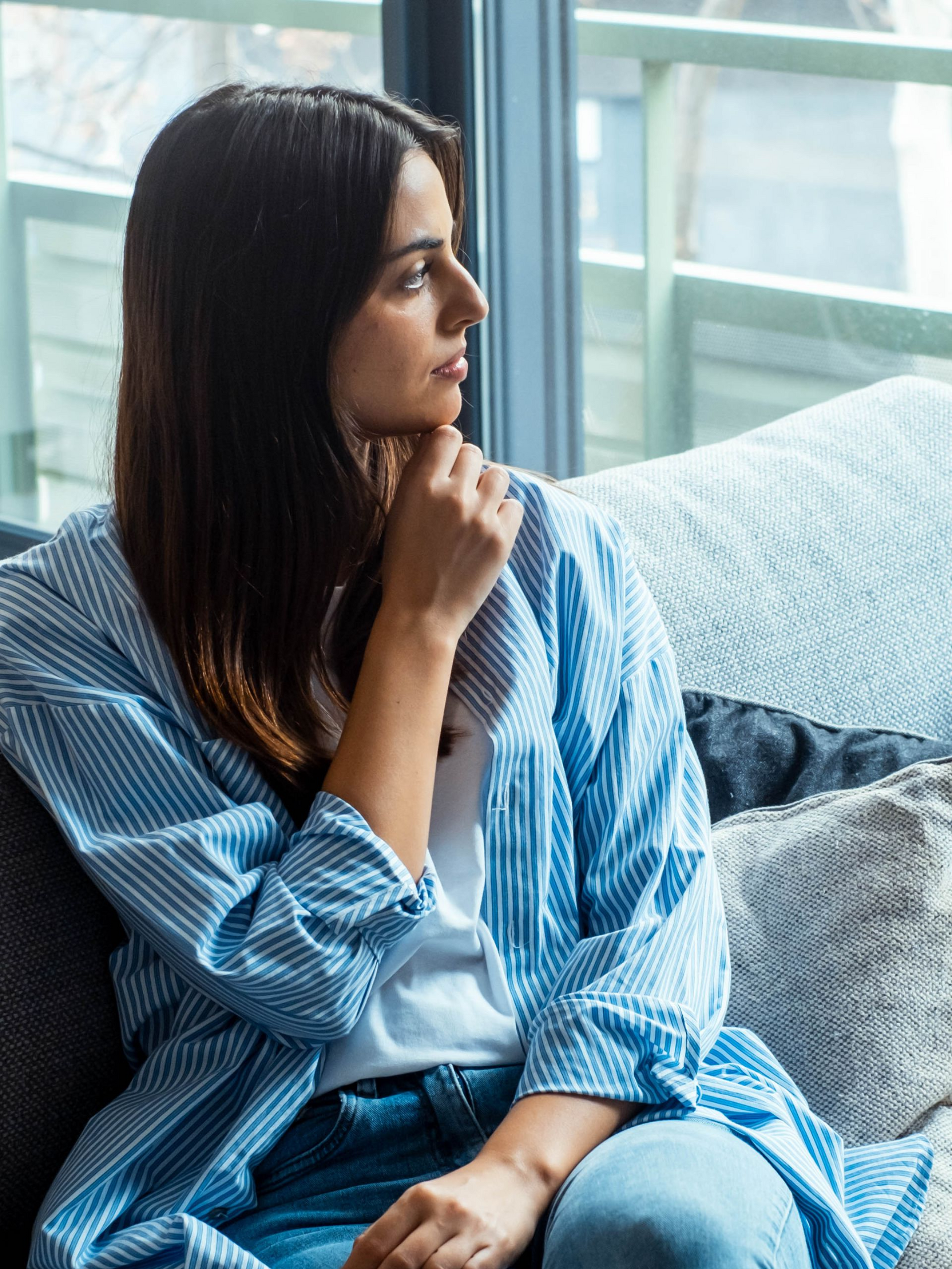 Woman in blue striped shirt gazes out window, resting chin on hand, seated on couch.