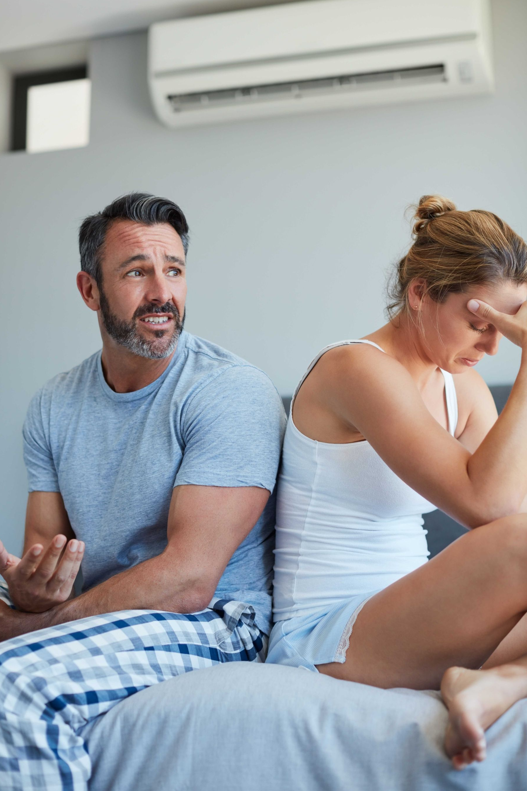 Man and woman sitting on bed, back to back. Both appear upset. Gray walls, air conditioner.