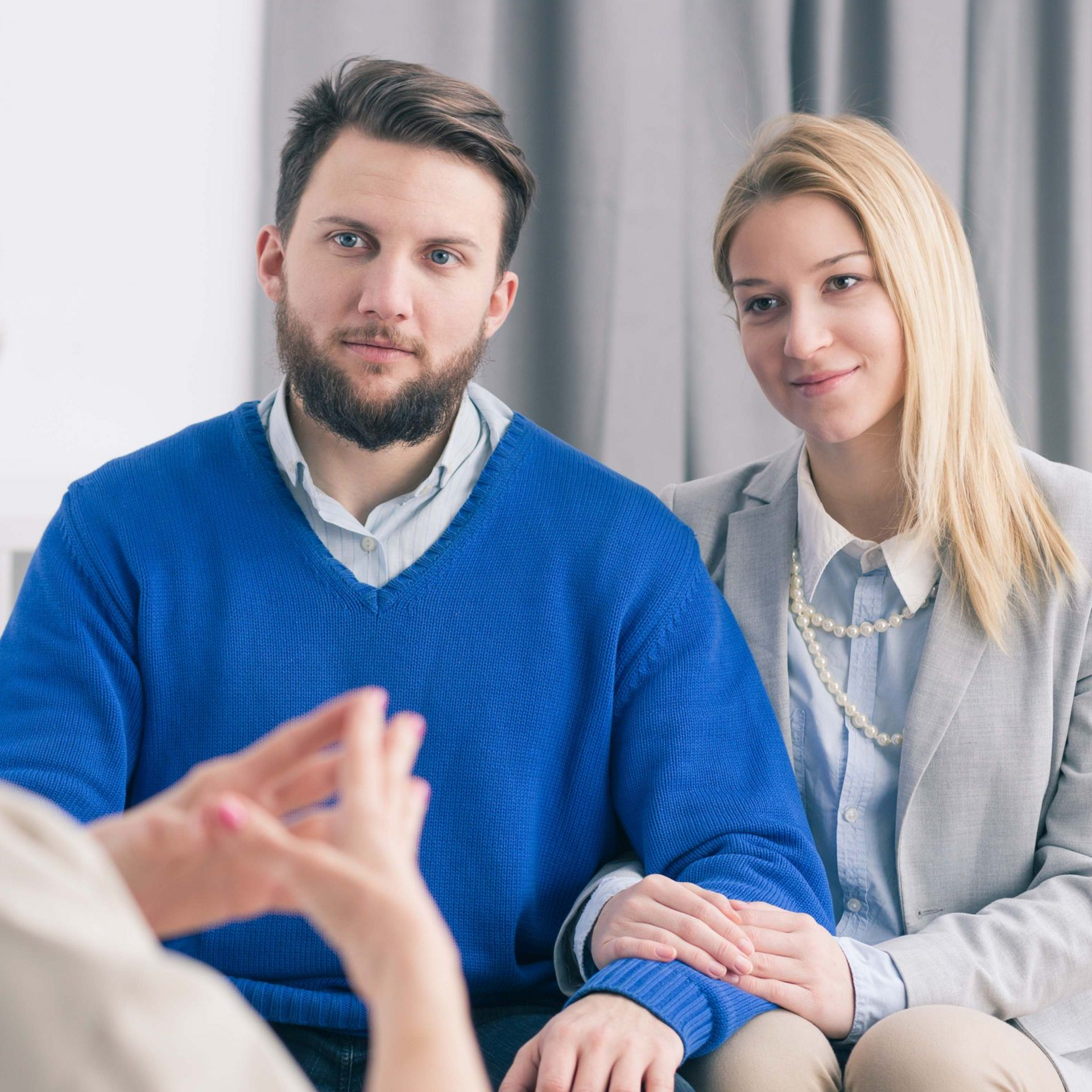 Couple in therapy session, man in blue sweater, woman in gray blazer, listening to therapist.