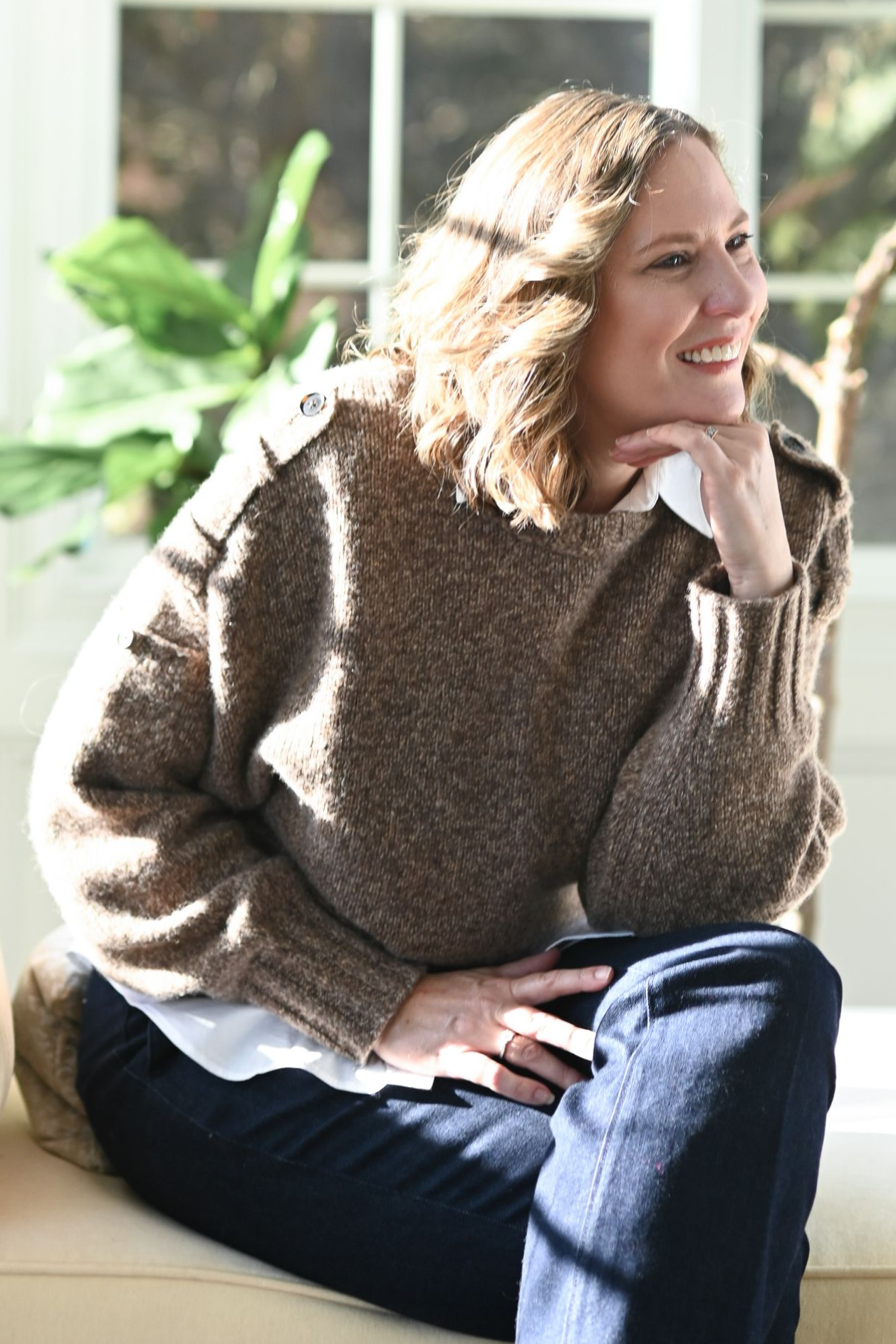 Woman smiling, wearing a burgundy leather jacket and cream-colored blouse, standing in front of a stone wall.