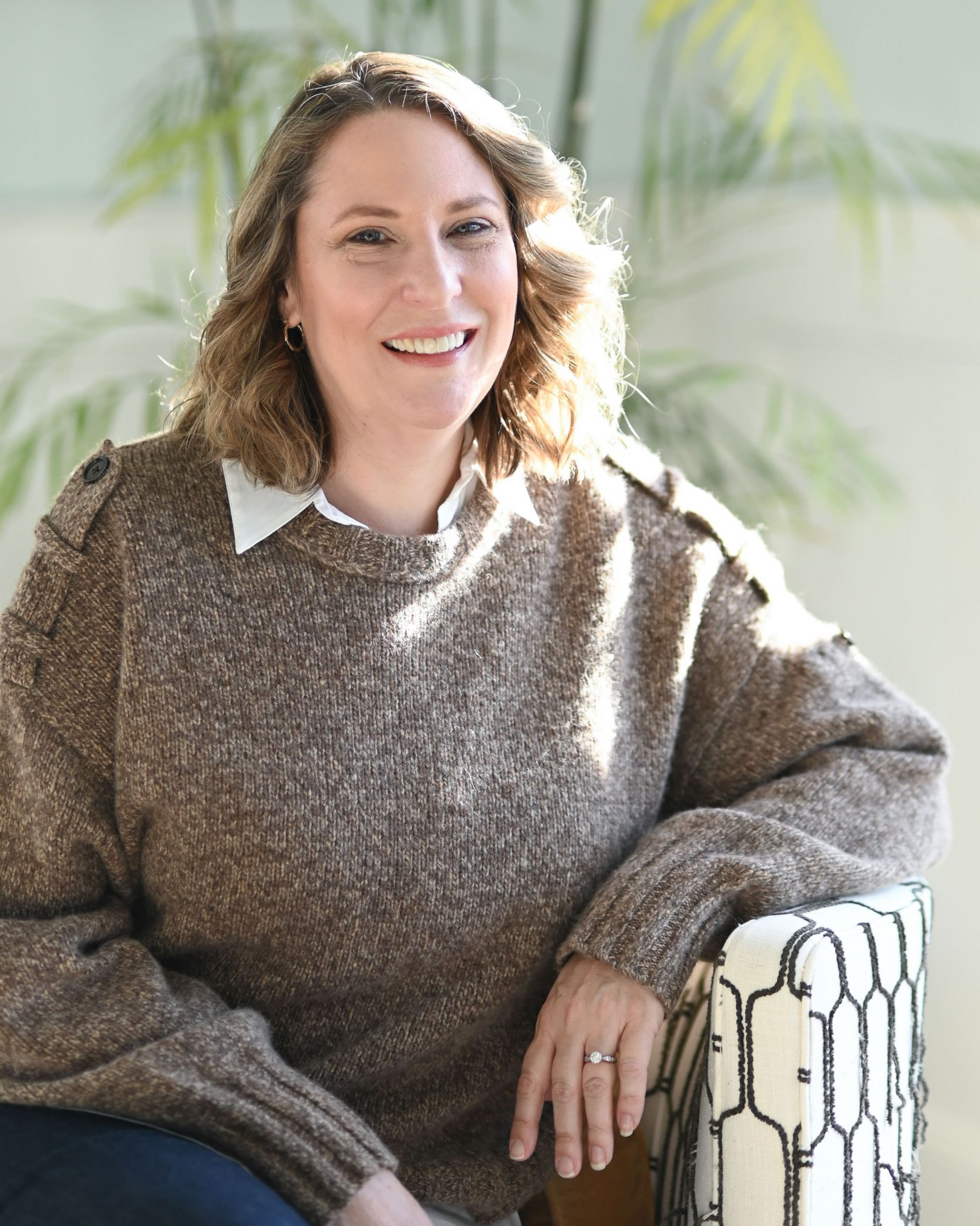 Woman smiling, wearing a burgundy leather jacket and cream-colored blouse, standing in front of a stone wall.