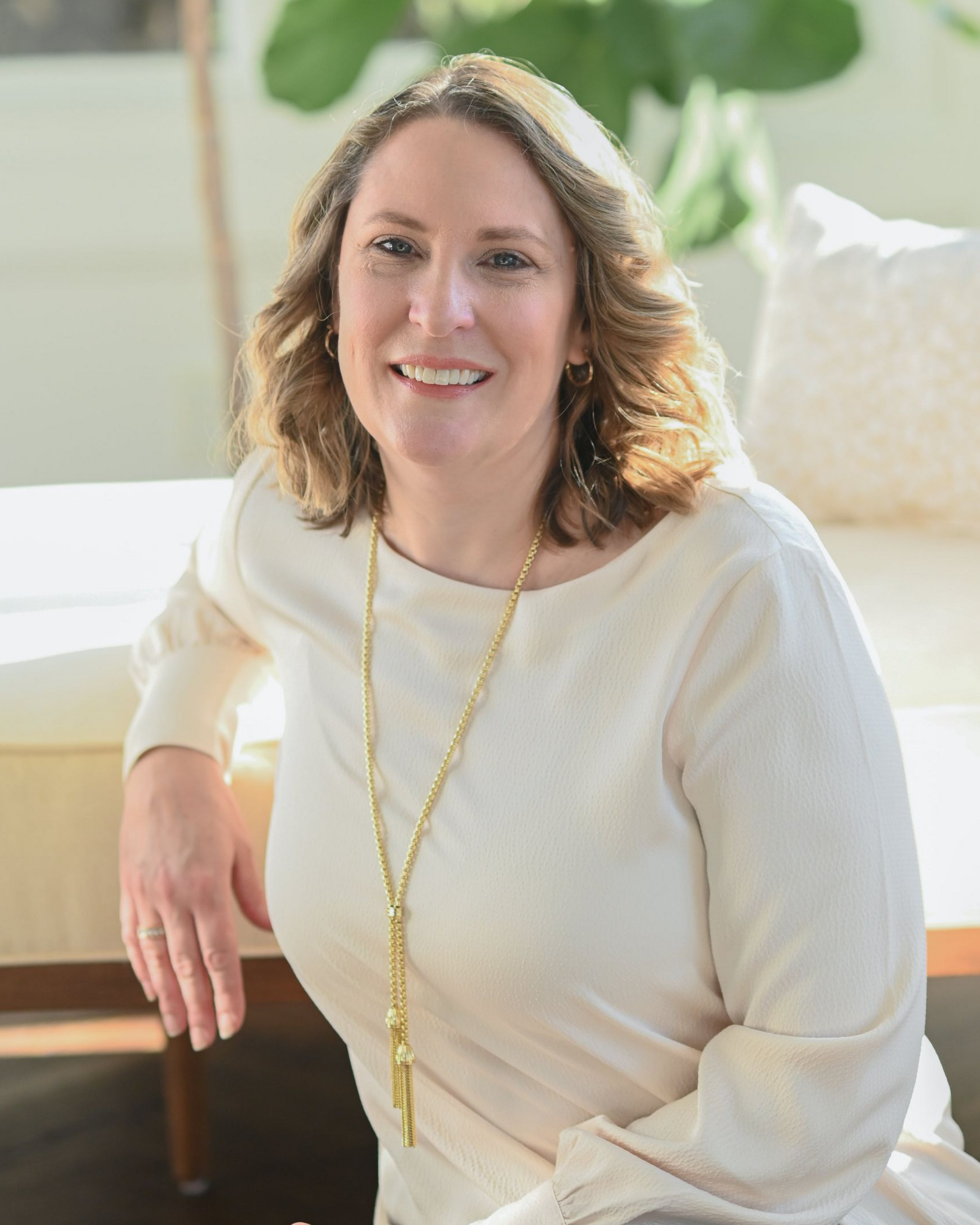Woman smiling, wearing a burgundy leather jacket and cream-colored blouse, standing in front of a stone wall.