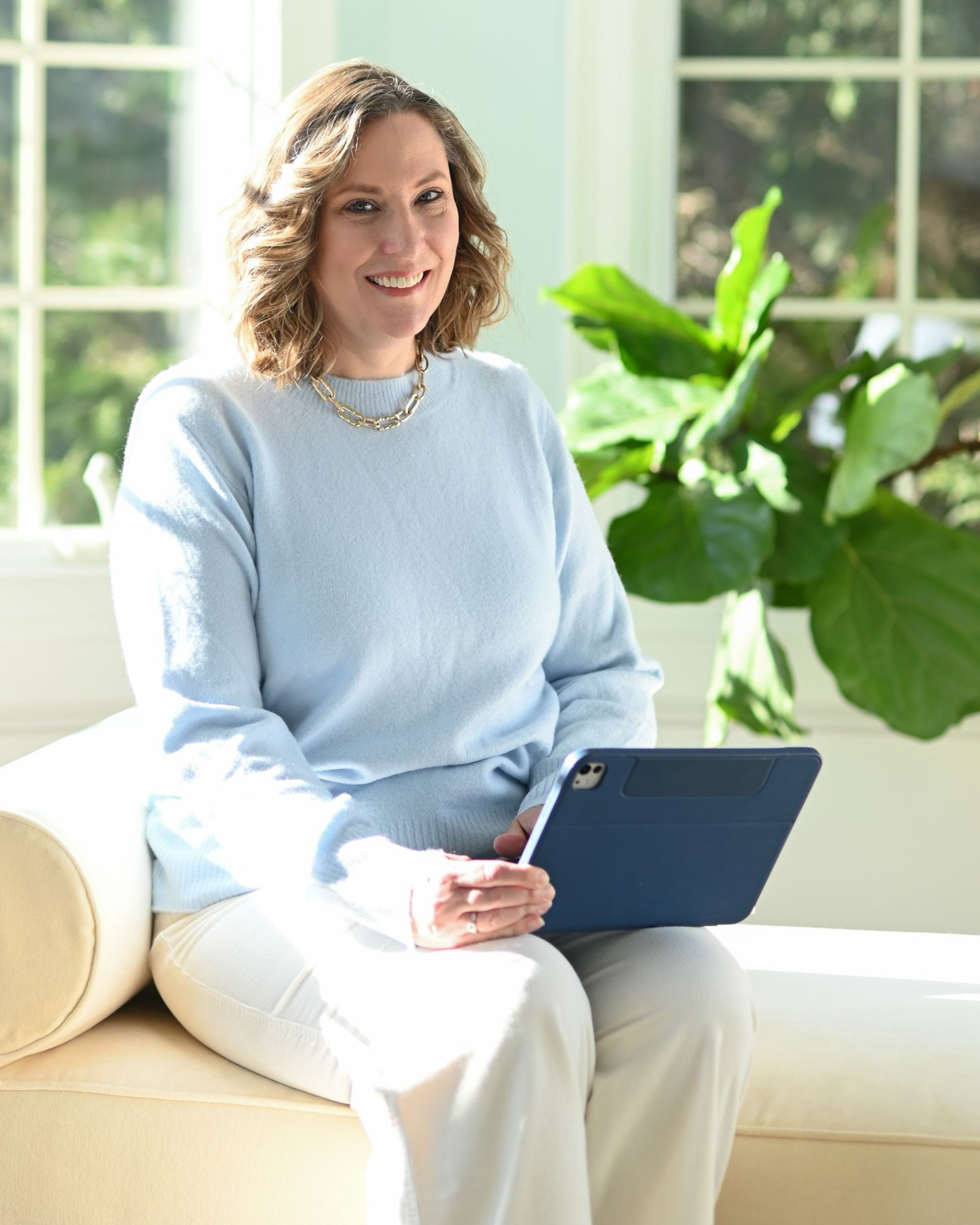 Woman at wooden table, typing on laptop, smiling. Man on green sofa in background.