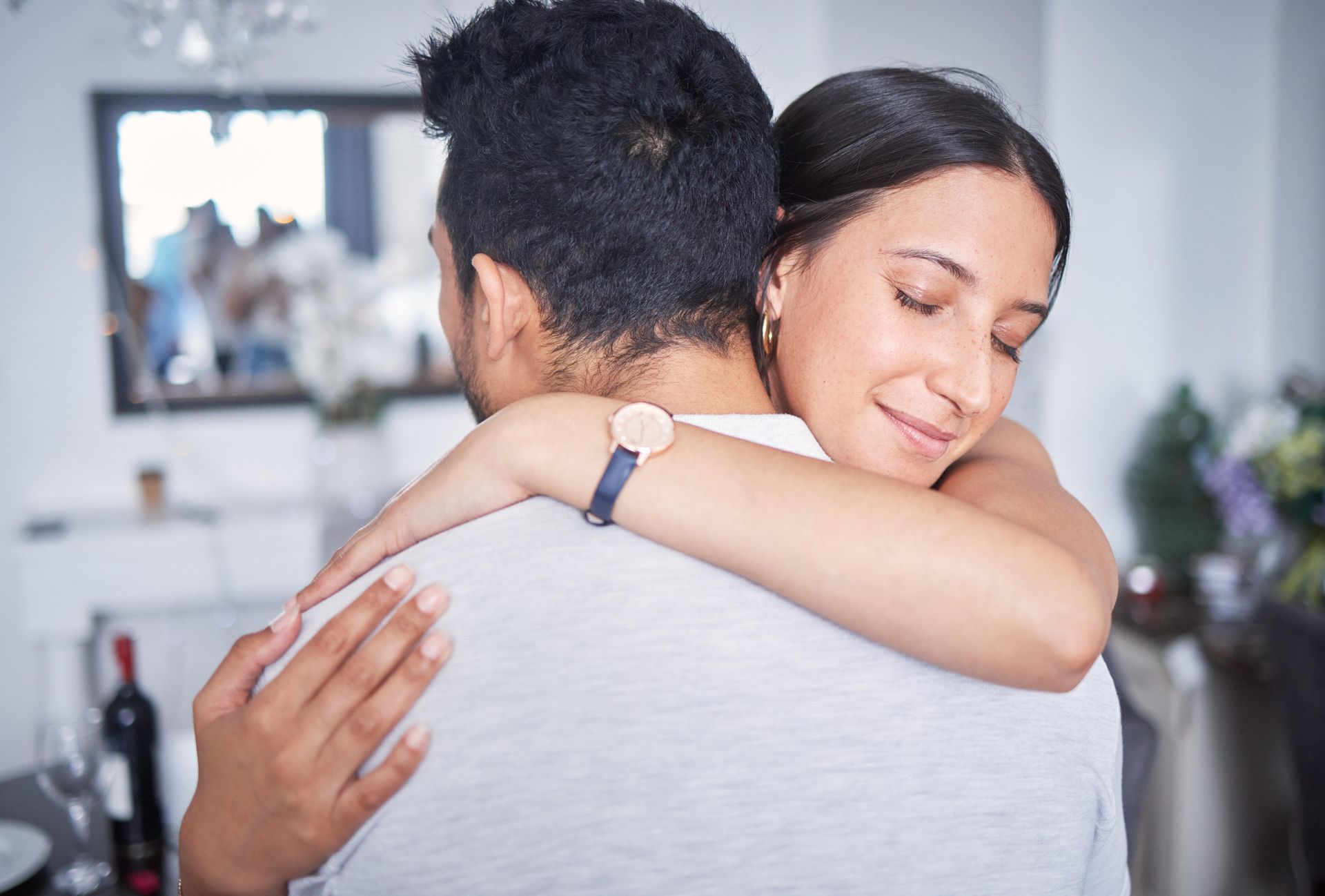 Woman with eyes closed hugs a person; a living room with a mirror and flowers is the background.
