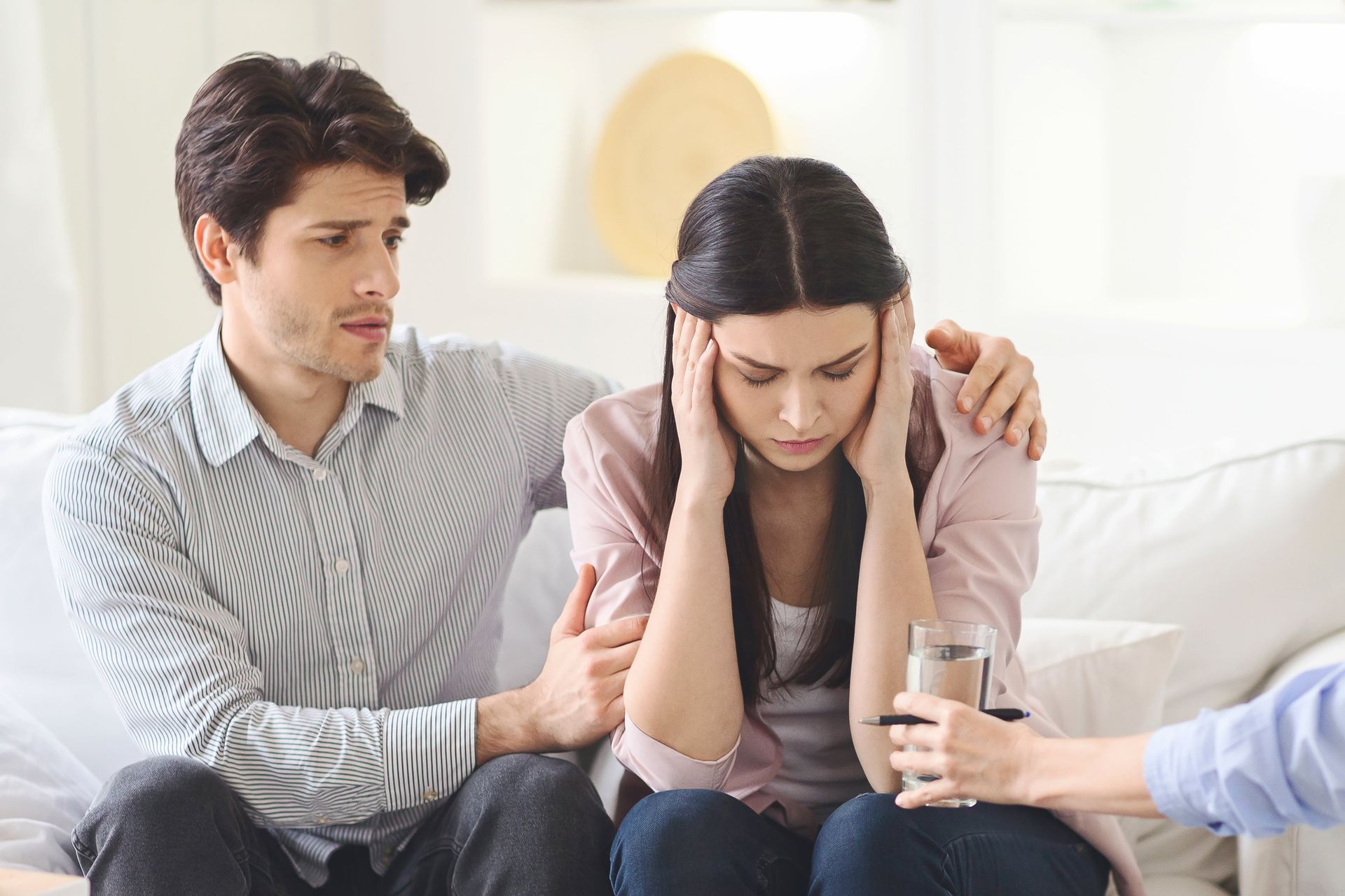 Couple in therapy, woman with hands on head, man consoles her. Therapist offers water.